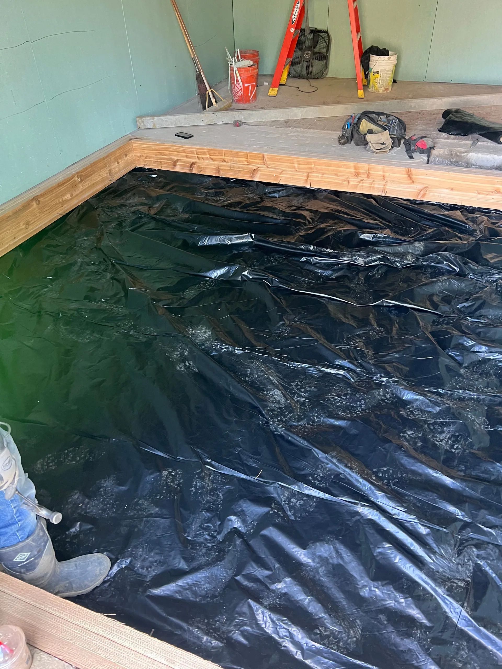 A construction site showing a black vapor barrier installed over floor joists, with drywall and tools visible in back.