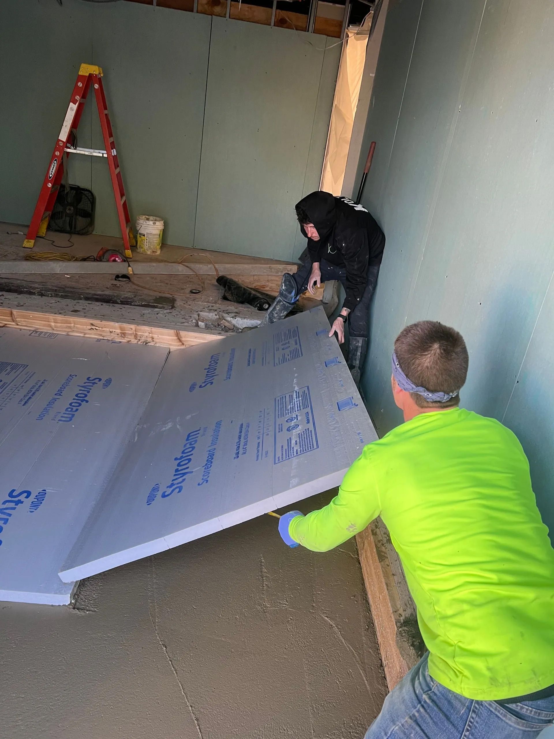 Two people in a room under construction lay large gray insulation panels on the concrete floor.