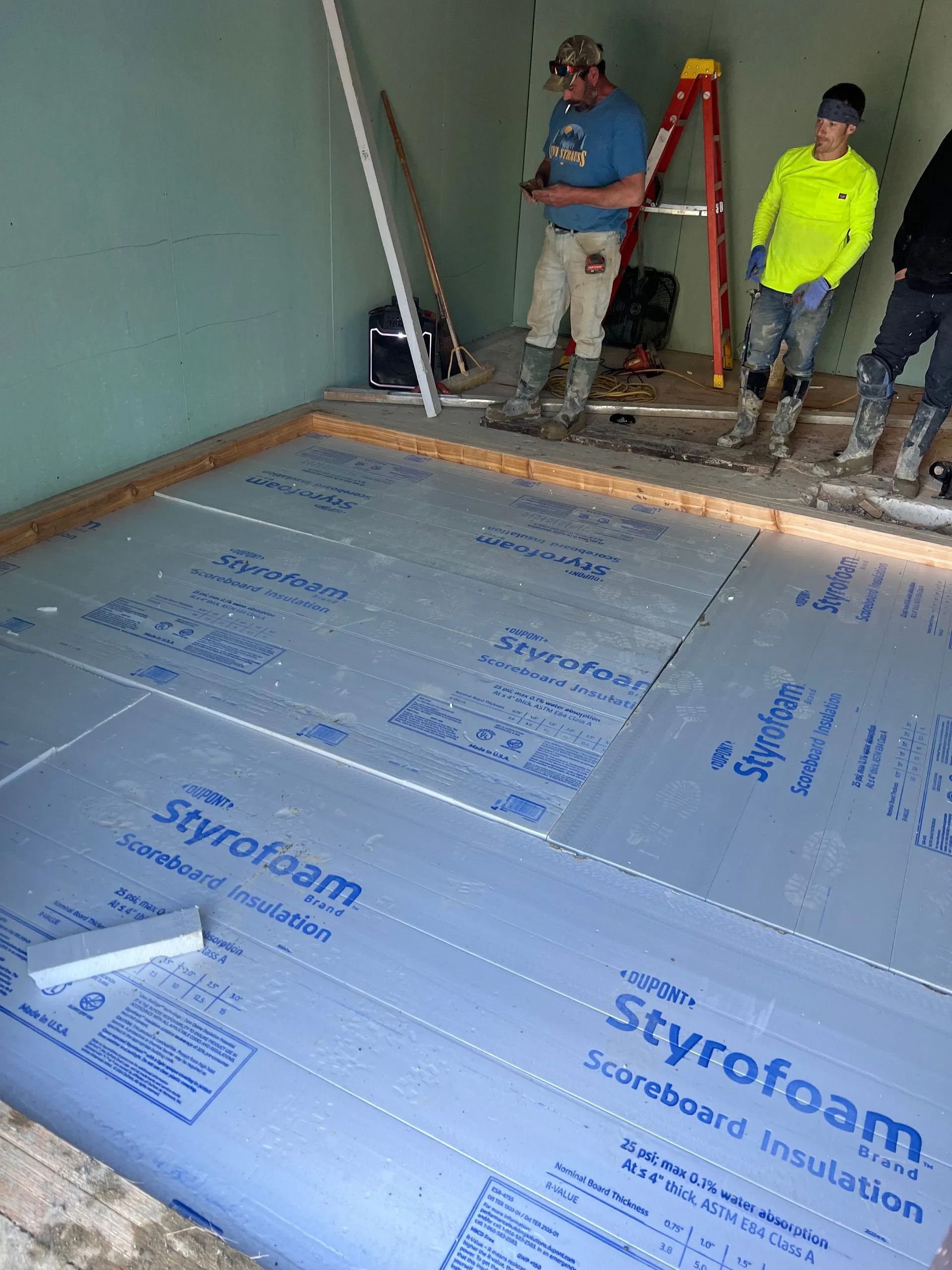 Two people work in a construction area, standing on a floor layered with blue Styrofoam insulation boards.