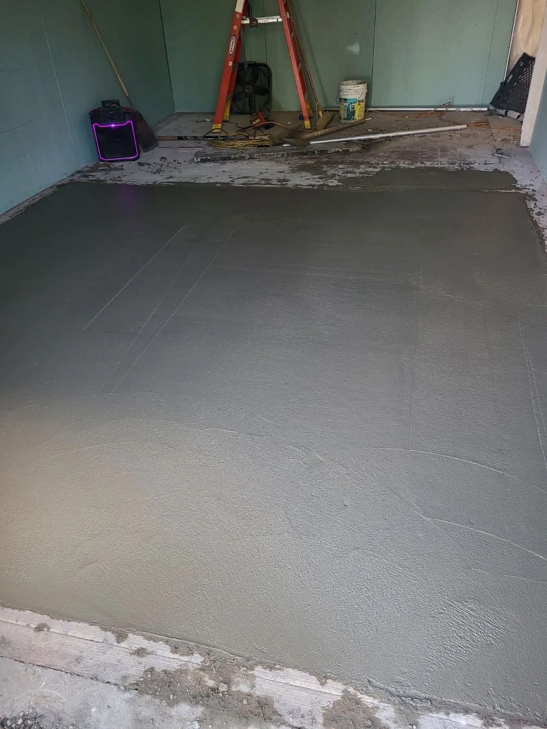 Freshly poured concrete floor in an unfinished room with a ladder and construction supplies in the background.