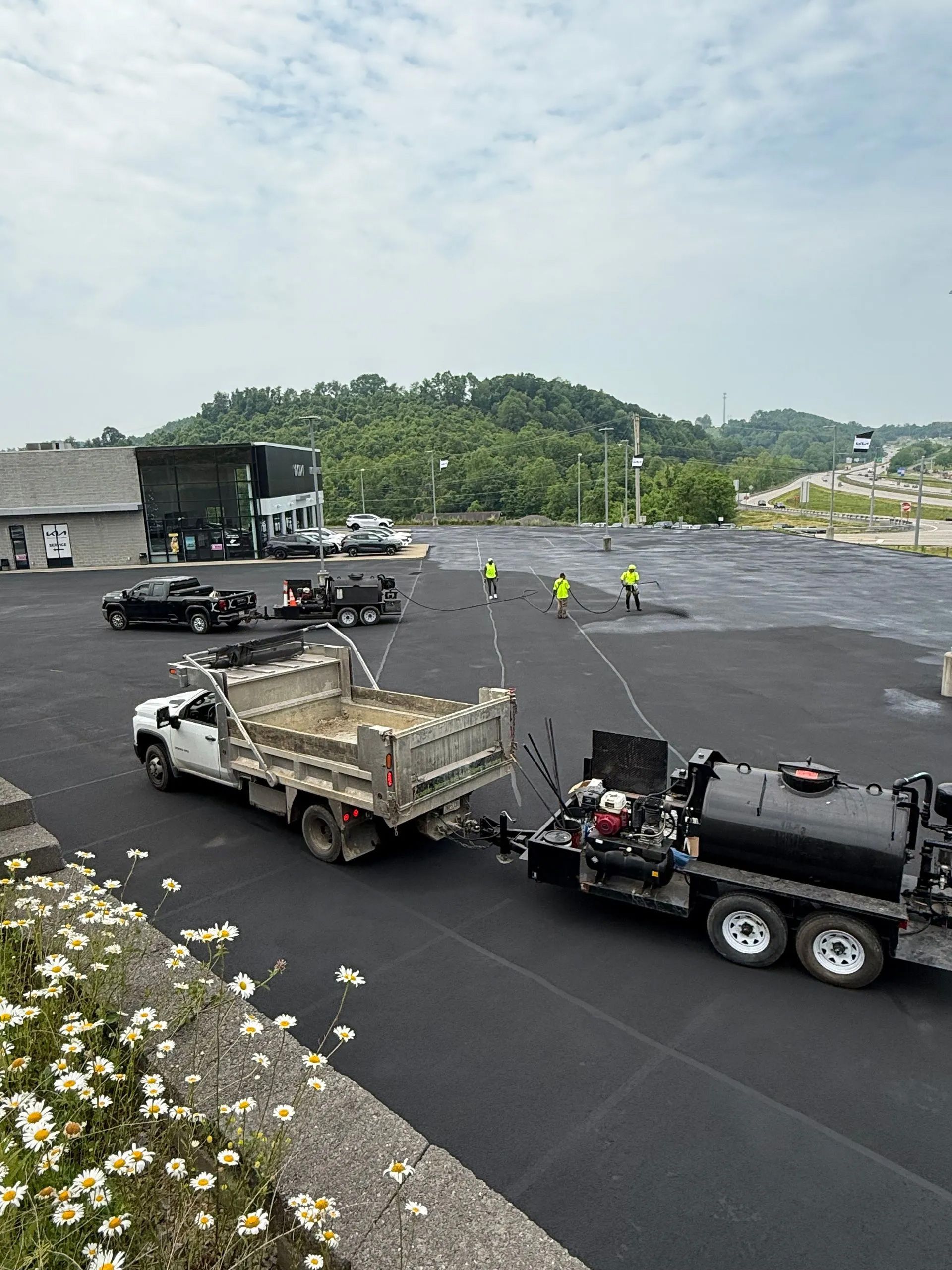 A pickup truck towing a spray trailer on a large, freshly paved asphalt lot, with workers visible in the distance.