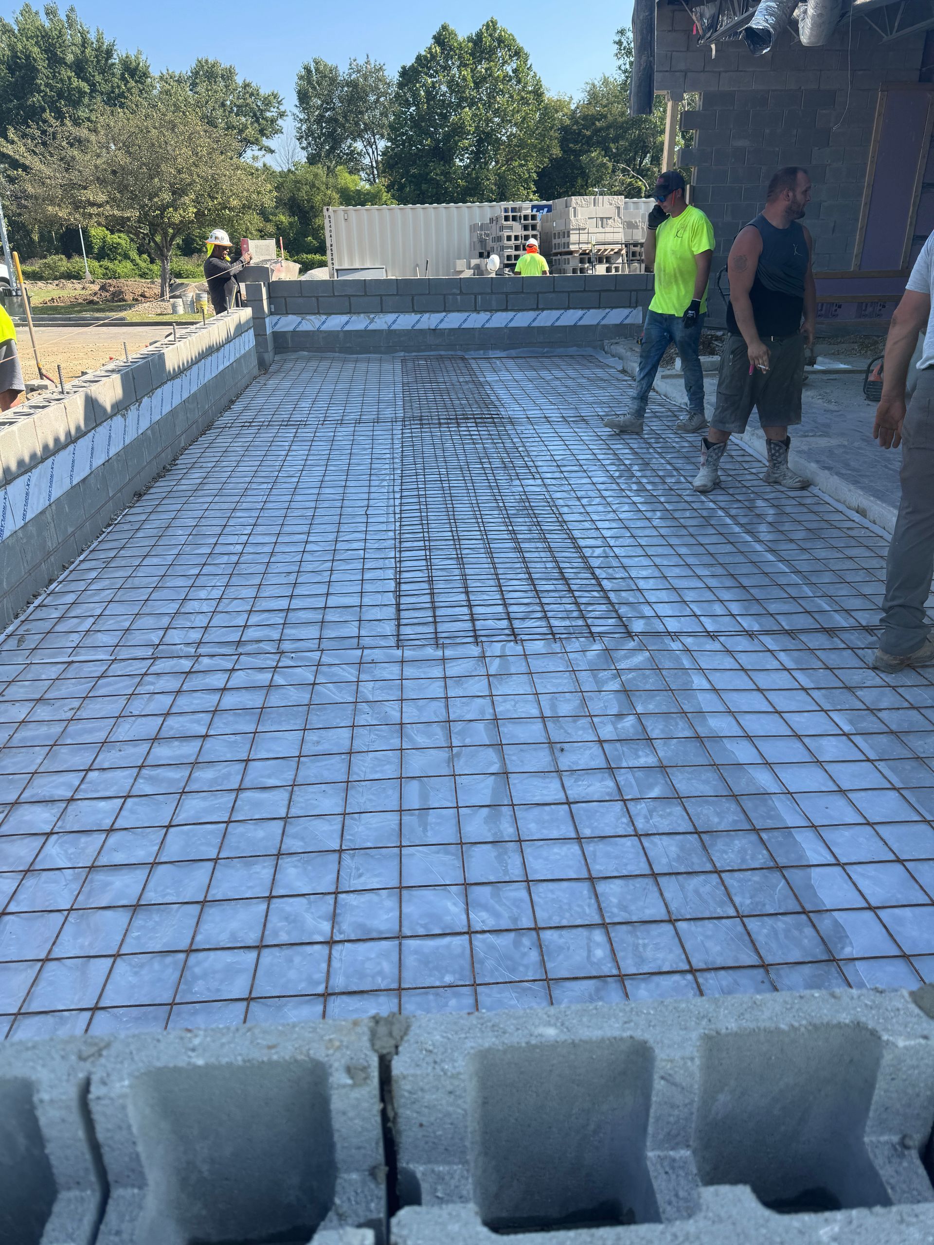 Construction workers prepare a rectangular area for concrete, featuring a metal grid laid over a reflective vapor barrier.