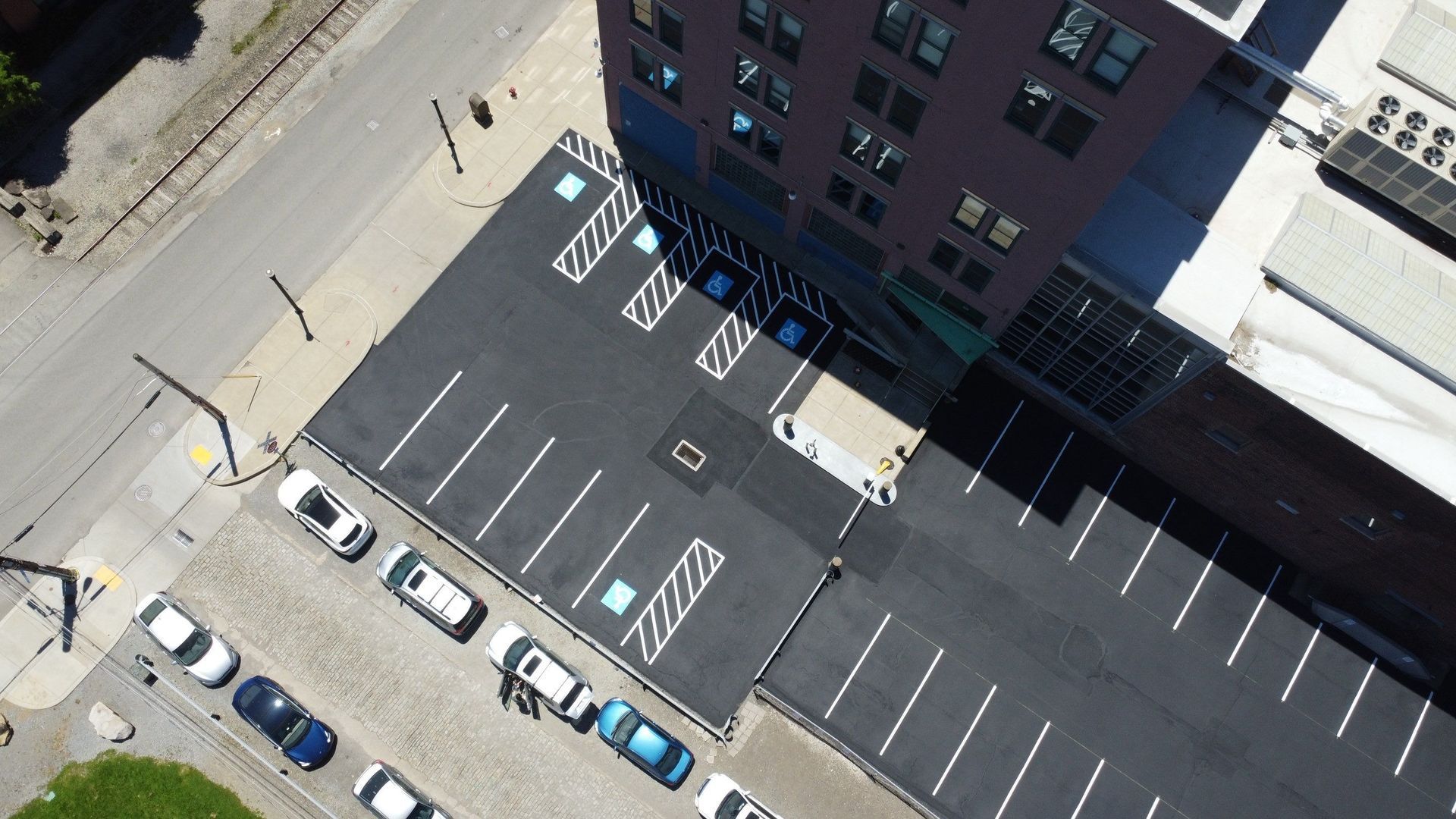 Aerial view of a mostly empty parking lot beside a building, with marked accessible spaces and parked cars along the street.