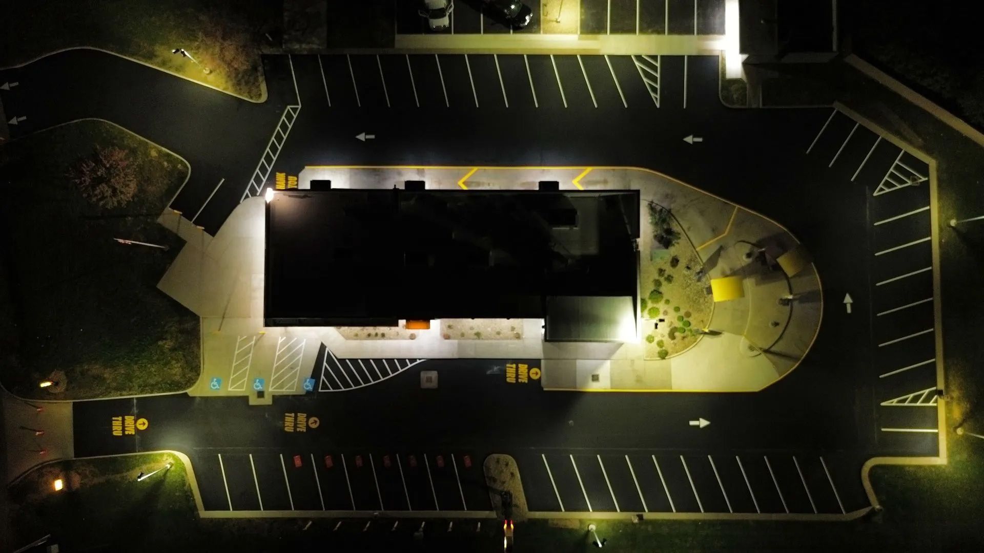 An aerial nighttime view of a brightly lit building surrounded by a parking lot with marked stalls and lanes.