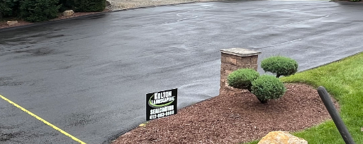 A freshly paved asphalt driveway with a stone pillar, landscaping, and a De-Jon Paving company sign.