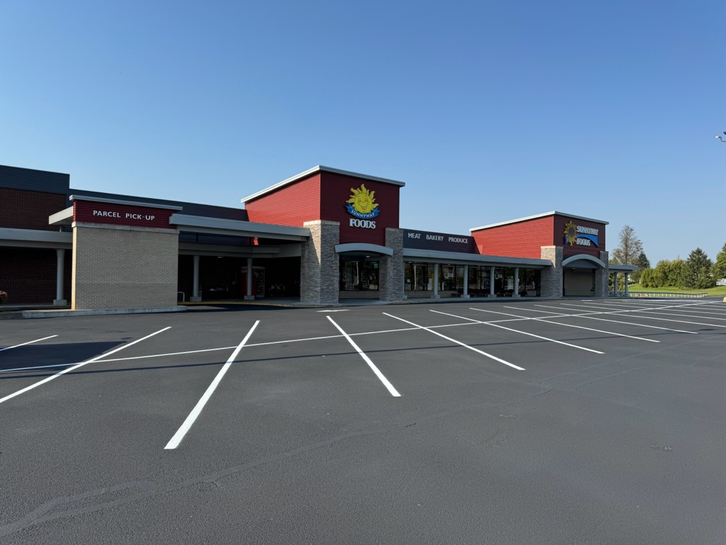 An empty parking lot in front of a modern one-story retail strip mall with a red facade and a central yellow leaf logo.