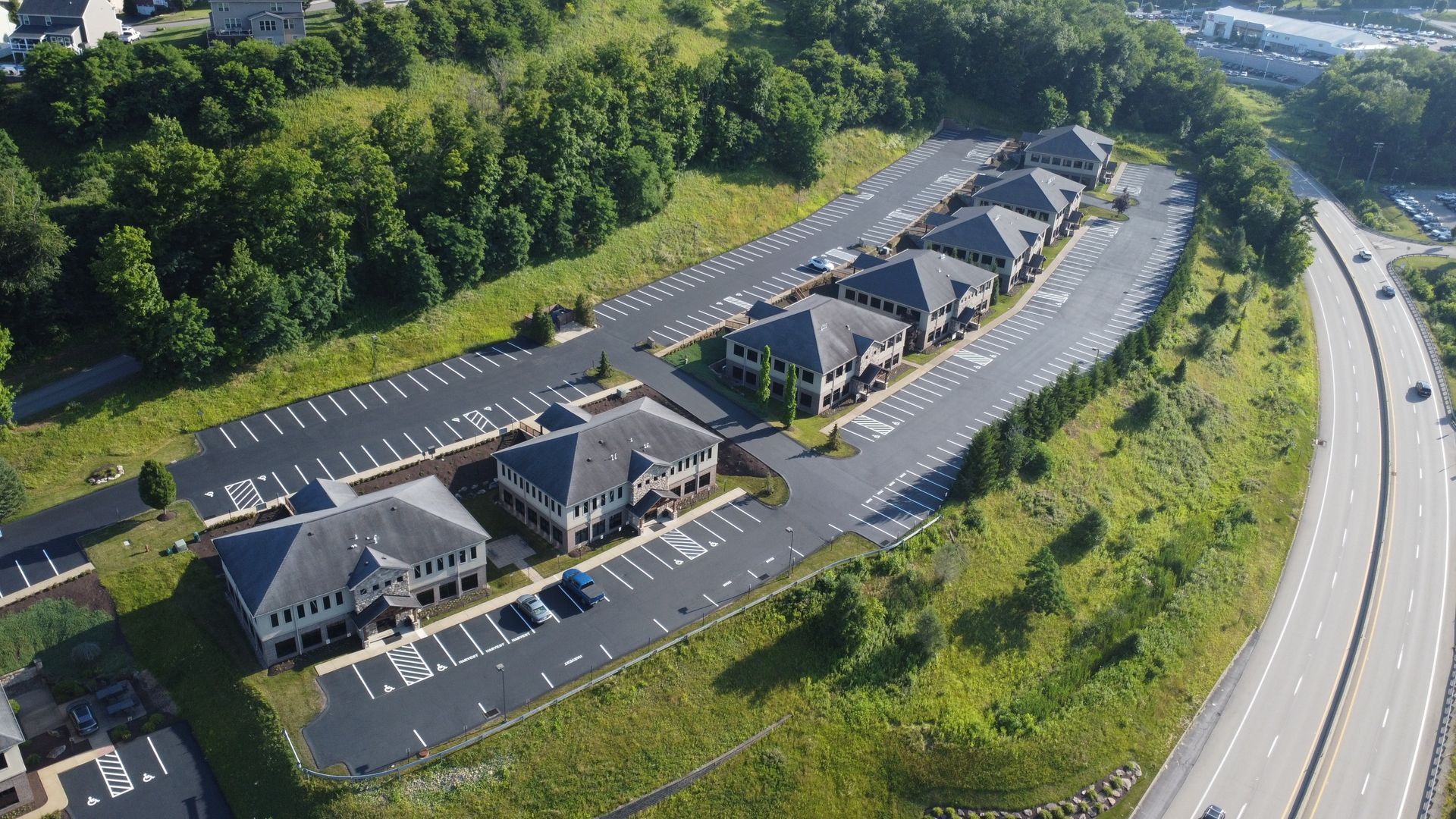Aerial view of a small commercial plaza with parking lots beside a highway and wooded hills.