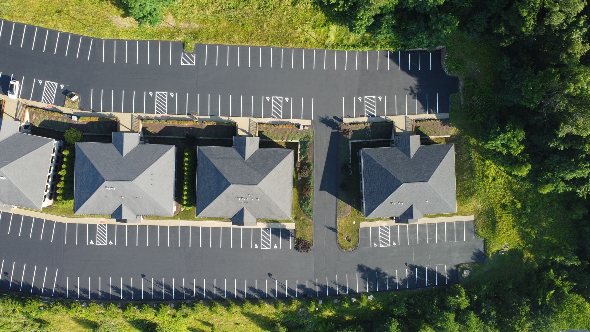 Aerial view of three gray-roofed houses in a mostly empty parking lot beside trees.