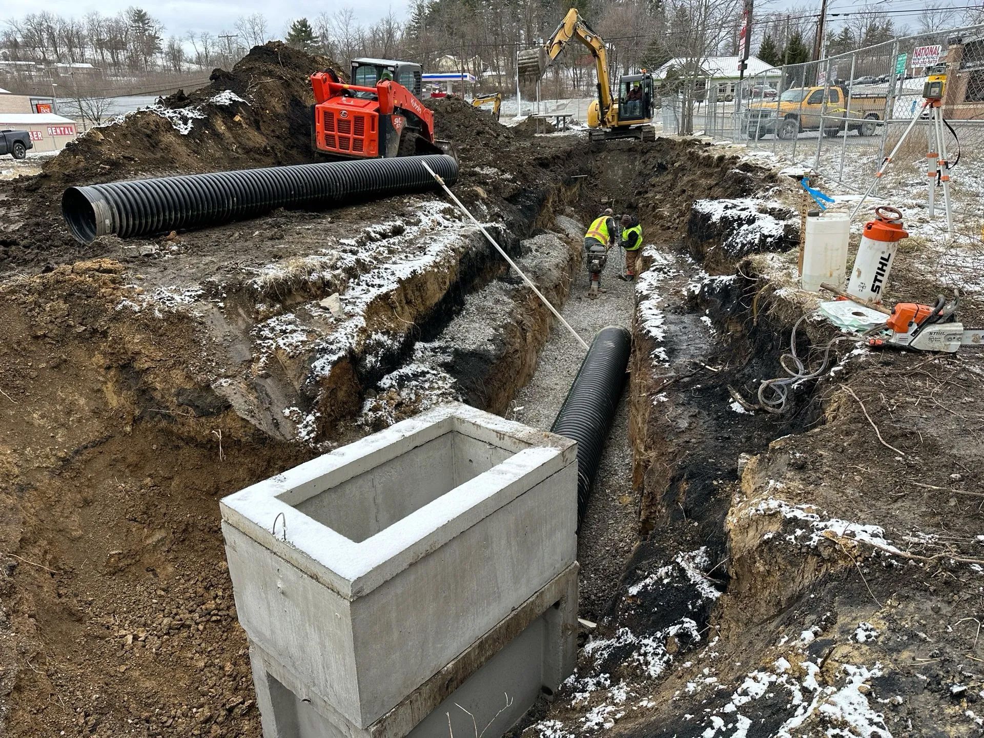 Construction site with an excavator and orange loader placing black drainage pipes into a trench near a concrete inlet.