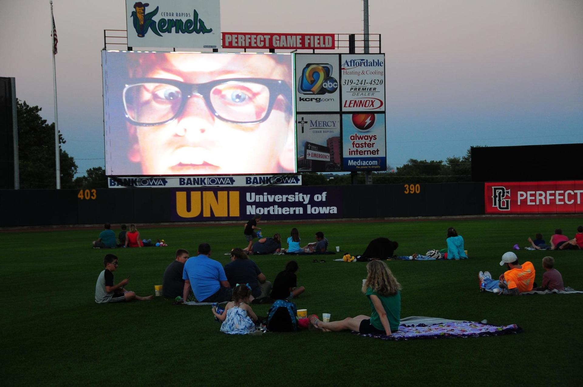 A group of people are sitting on the grass in front of a large screen that says university of northern iowa