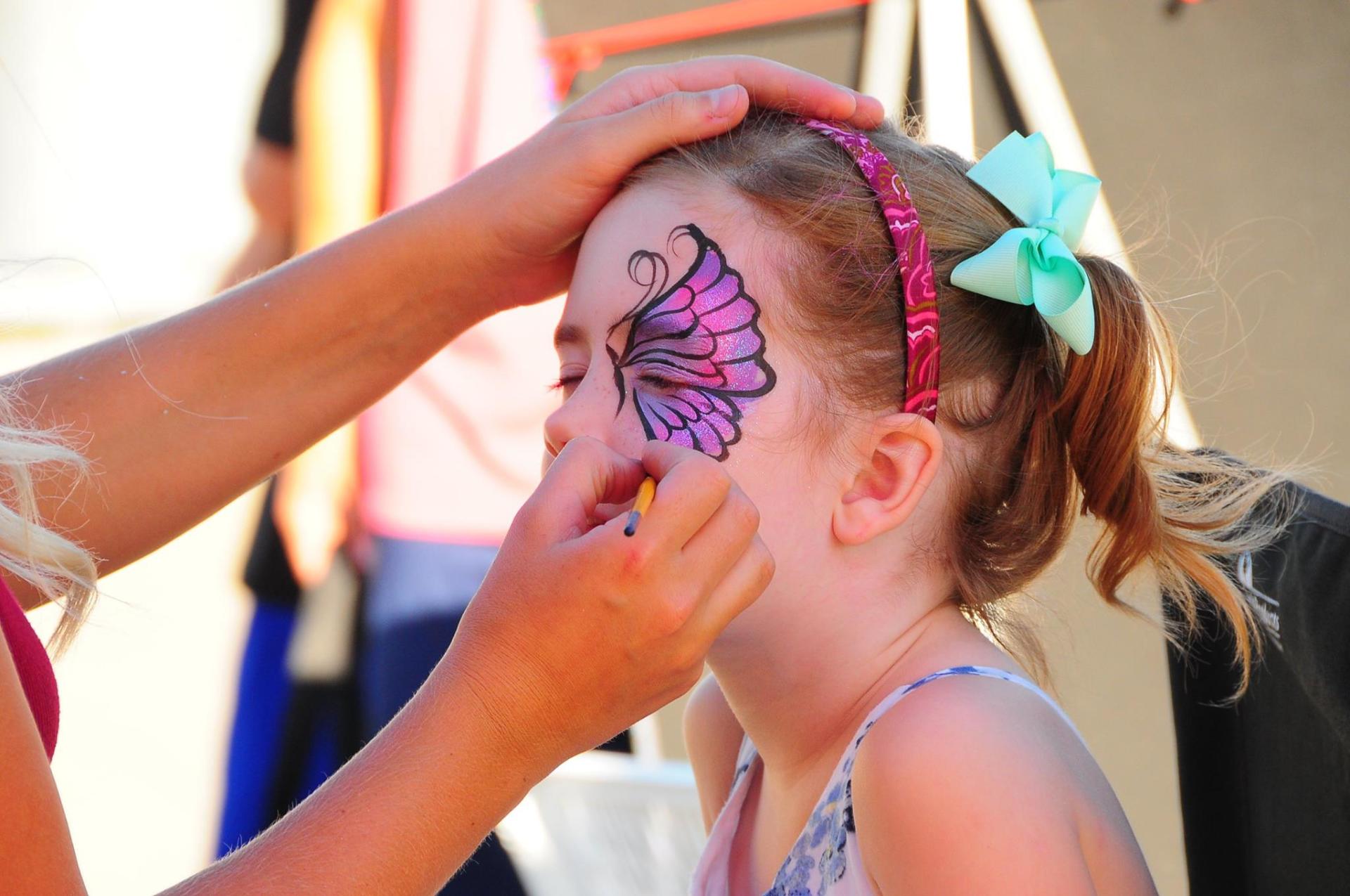 A little girl is getting her face painted with a butterfly.