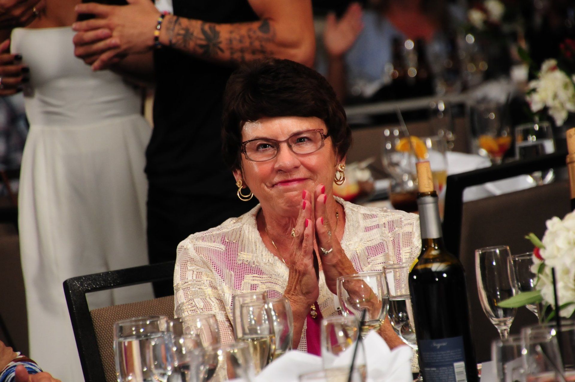 A woman is sitting at a table with glasses and a bottle of wine.