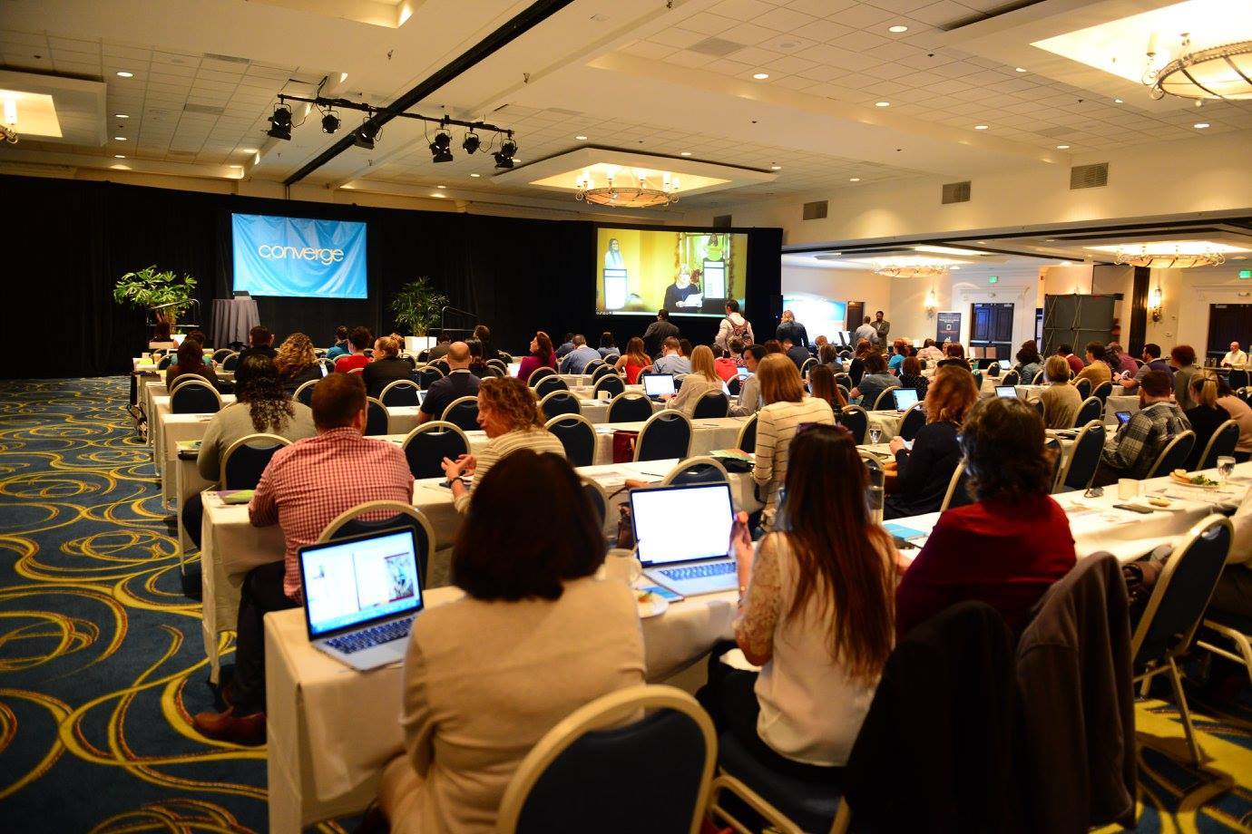A large group of people are sitting at tables with laptops in a conference room.