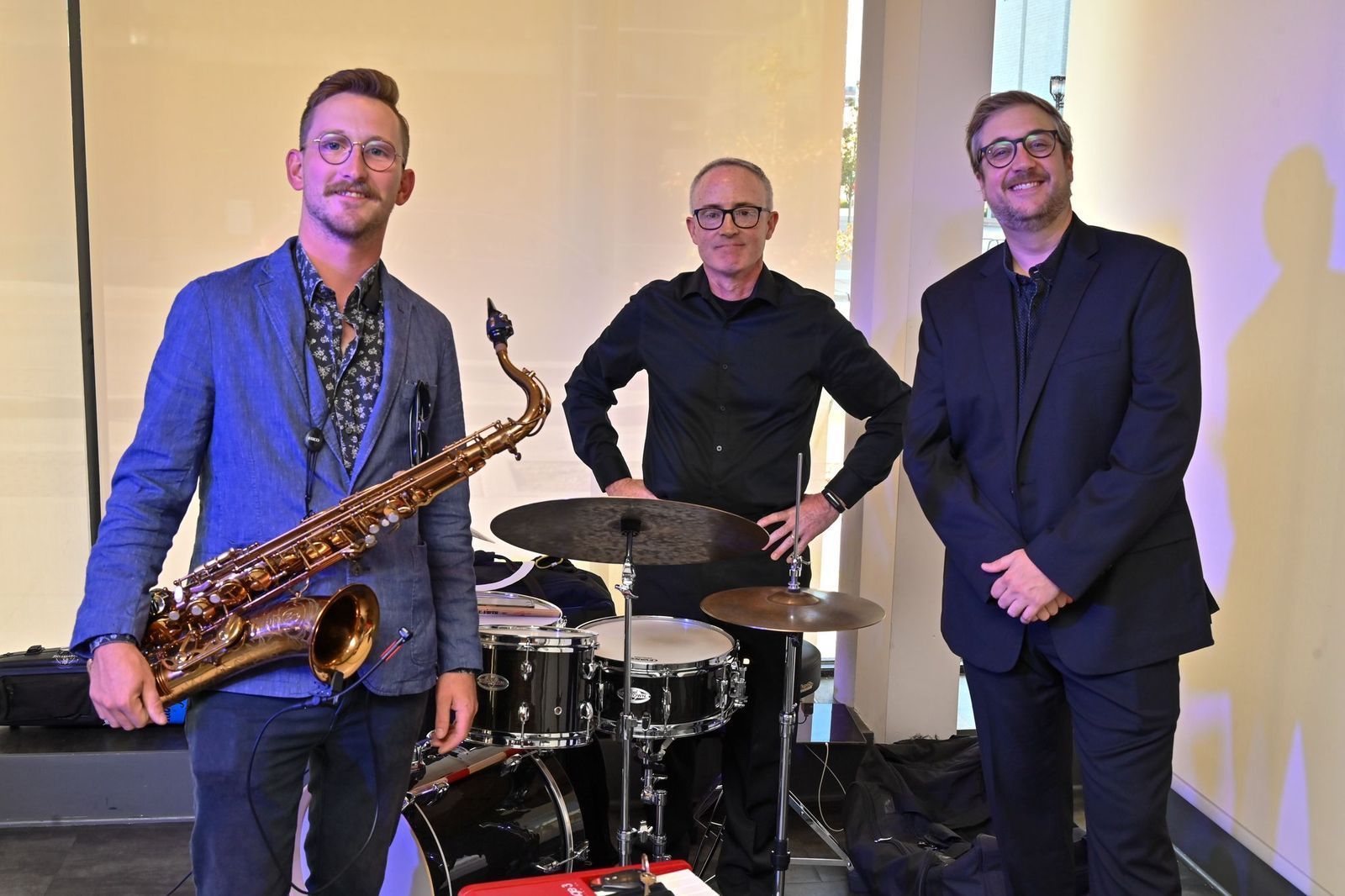 a guy holding a saxophone, a guy behind a drum set, and a guy standing wearing black