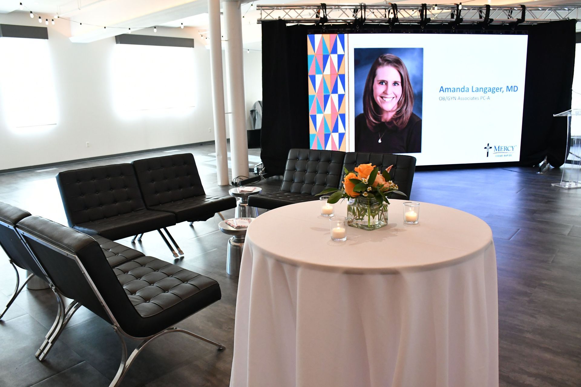 table with white cloth, flowers, and candles on top with black couches at the back and the speaker's information and photo flashed in the LED background