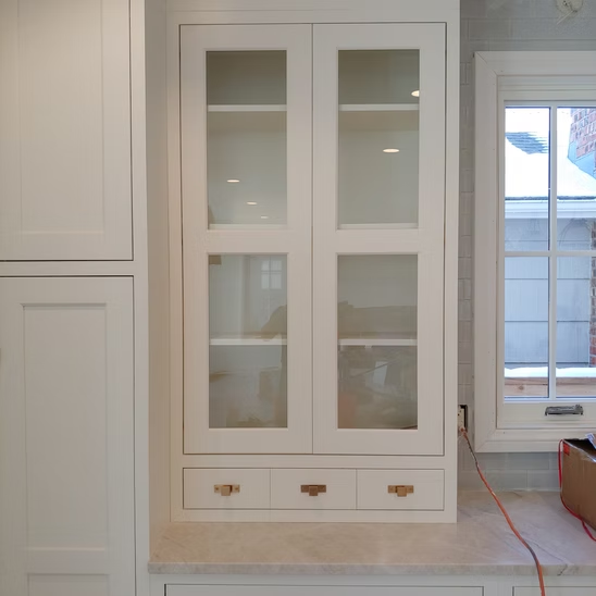 A white cabinet with glass doors and drawers in a kitchen next to a window.
