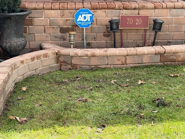 Brick home exterior with low brick retaining wall, grass, and security system sign after.