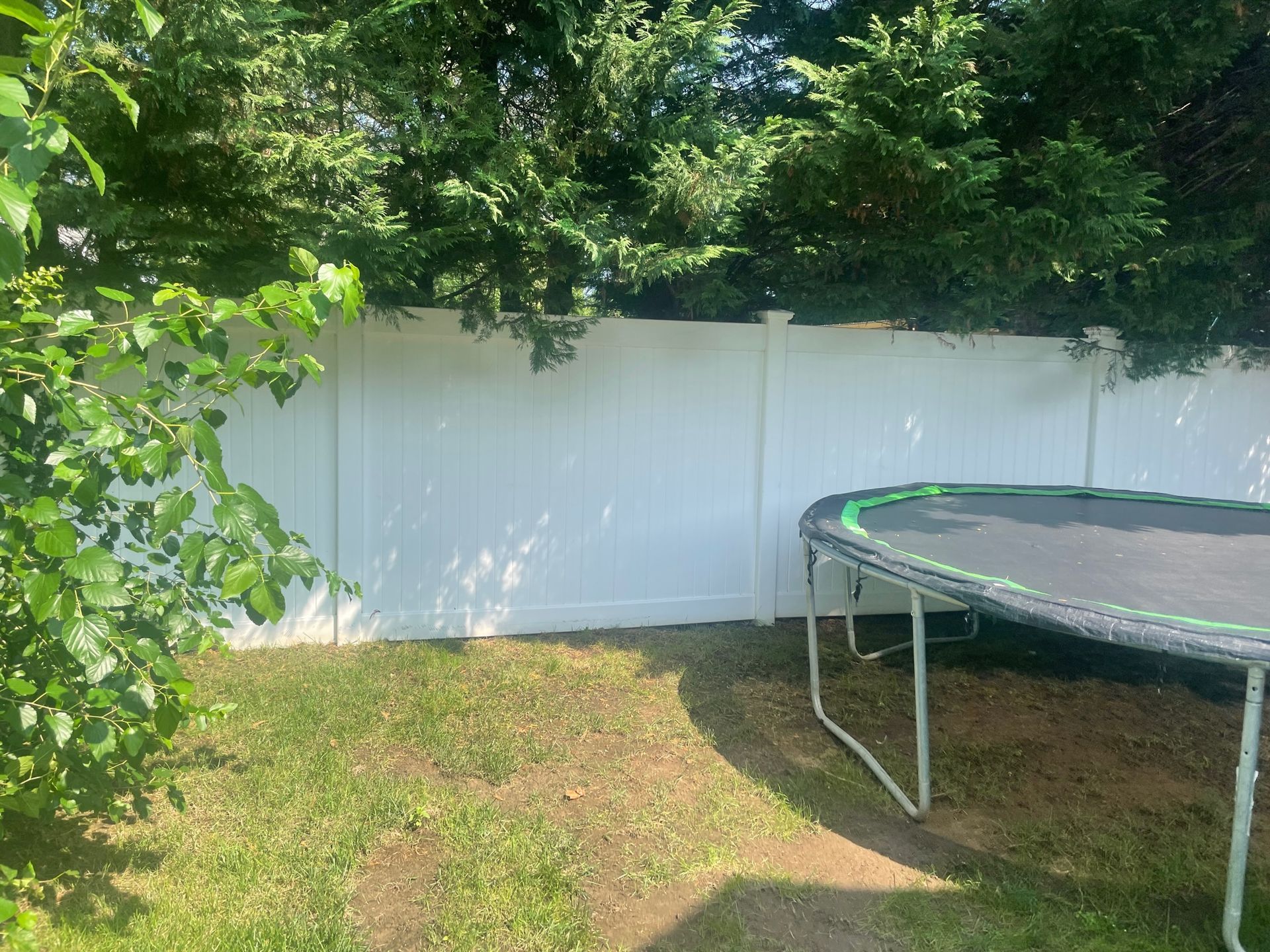 A backyard with a white fence, trampoline, and green grass. The fence has discoloration and some trees after.