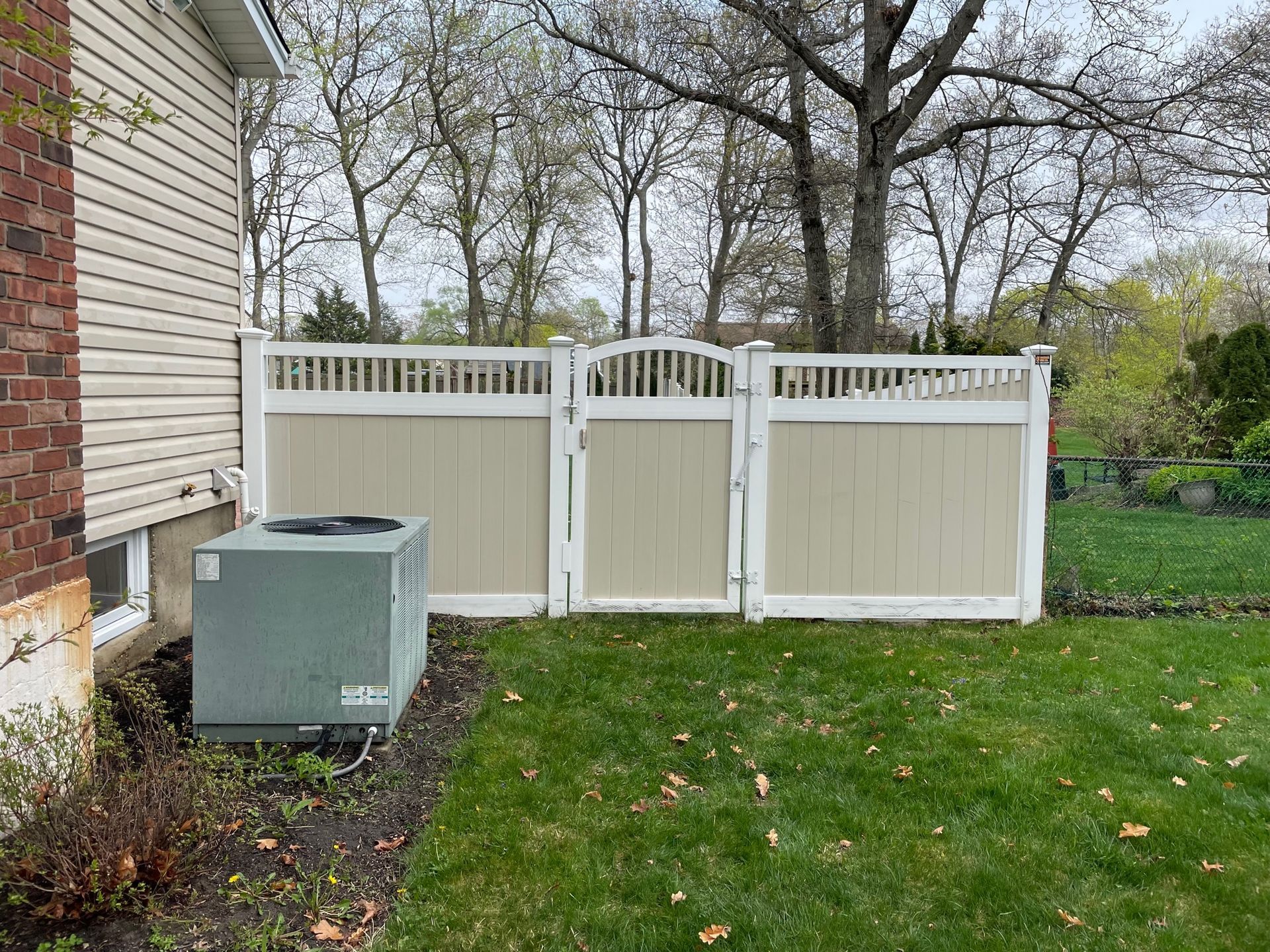 A beige vinyl fence in a backyard with a central gate next to an air conditioning unit after.