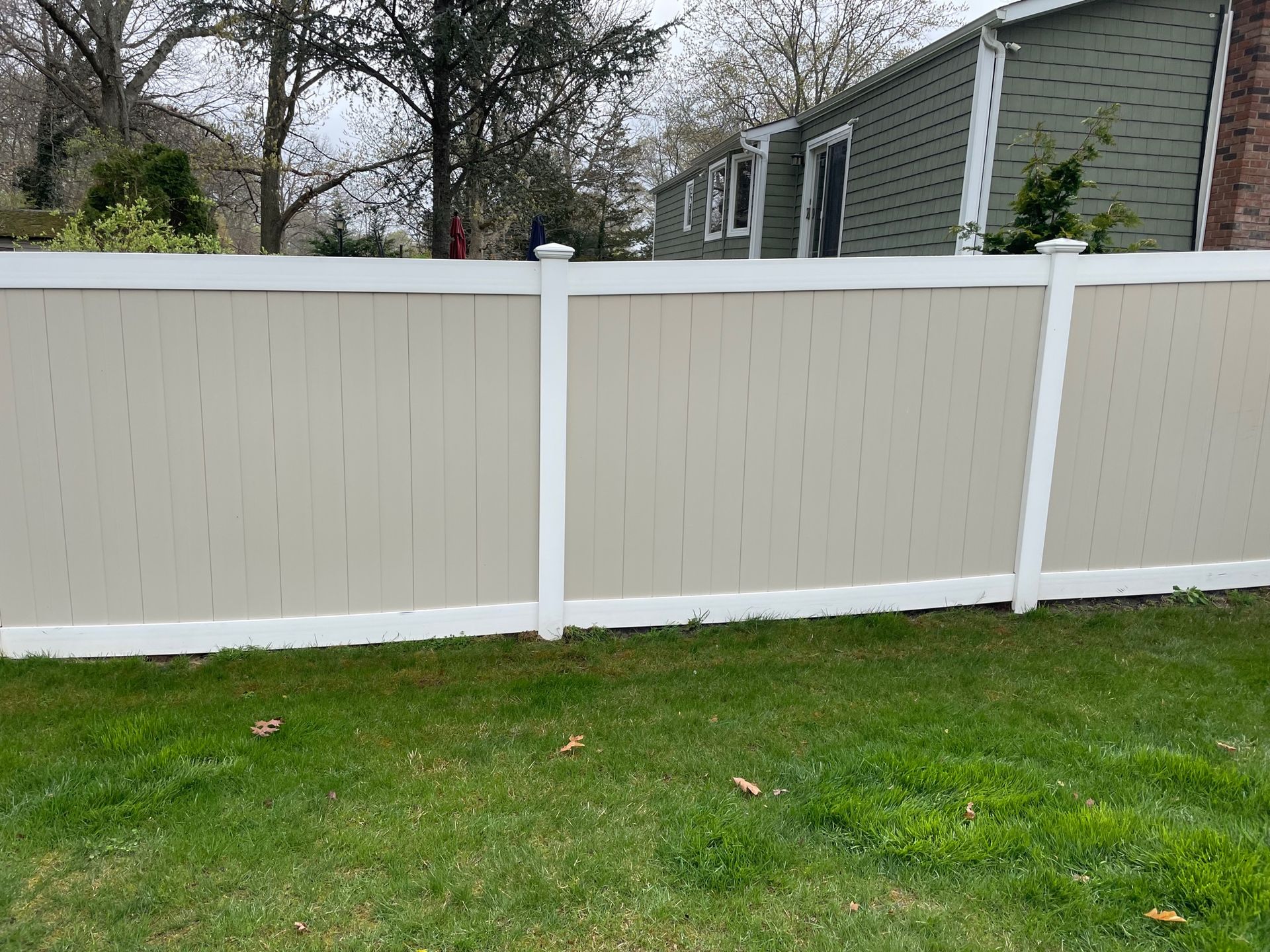 Tan vinyl fence with white posts, surrounded by grass, trees, and a house in the background after.