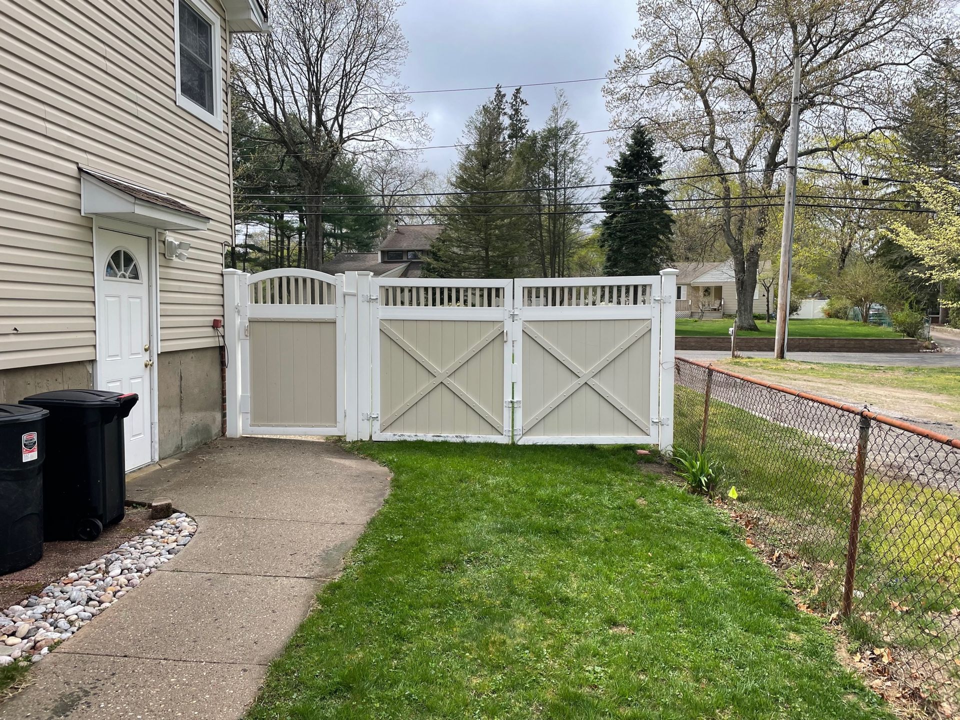 Back of a beige house with a white picket fence, two trash cans, and a grassy yard after.