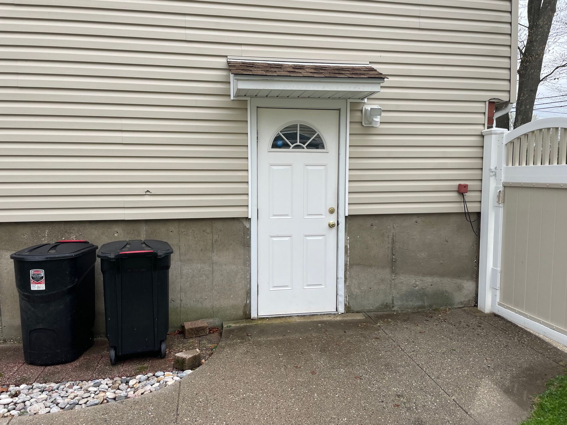 Back door of a house with a small awning, next to a white fence and trash cans after.