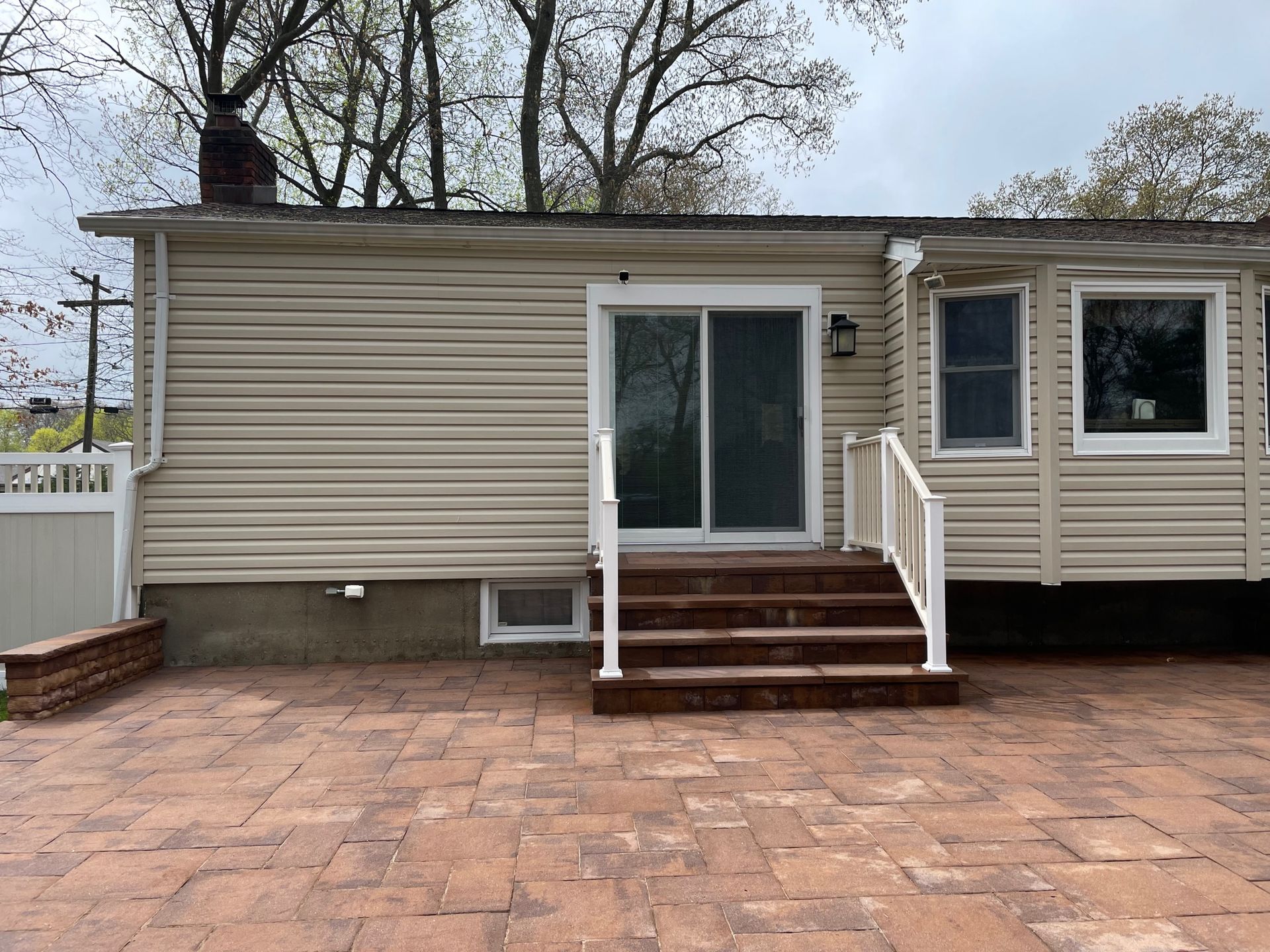 Back of house with brick patio, steps, and sliding door. Beige siding, white railing, overcast sky after.