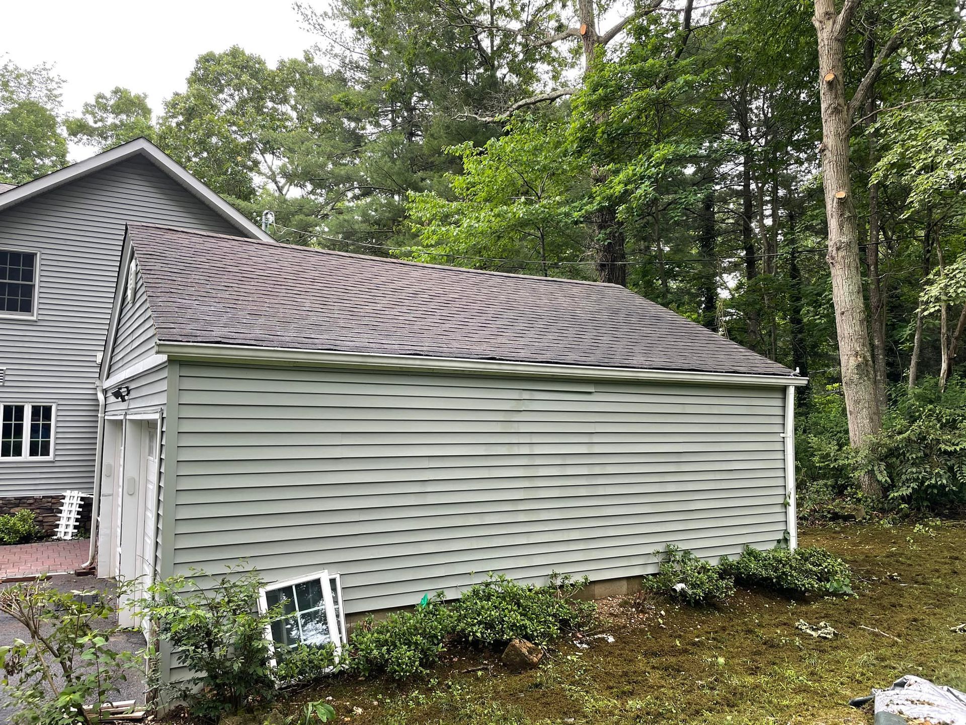 A light green building with a mossy roof being cleaned, surrounded by trees and bushes after.