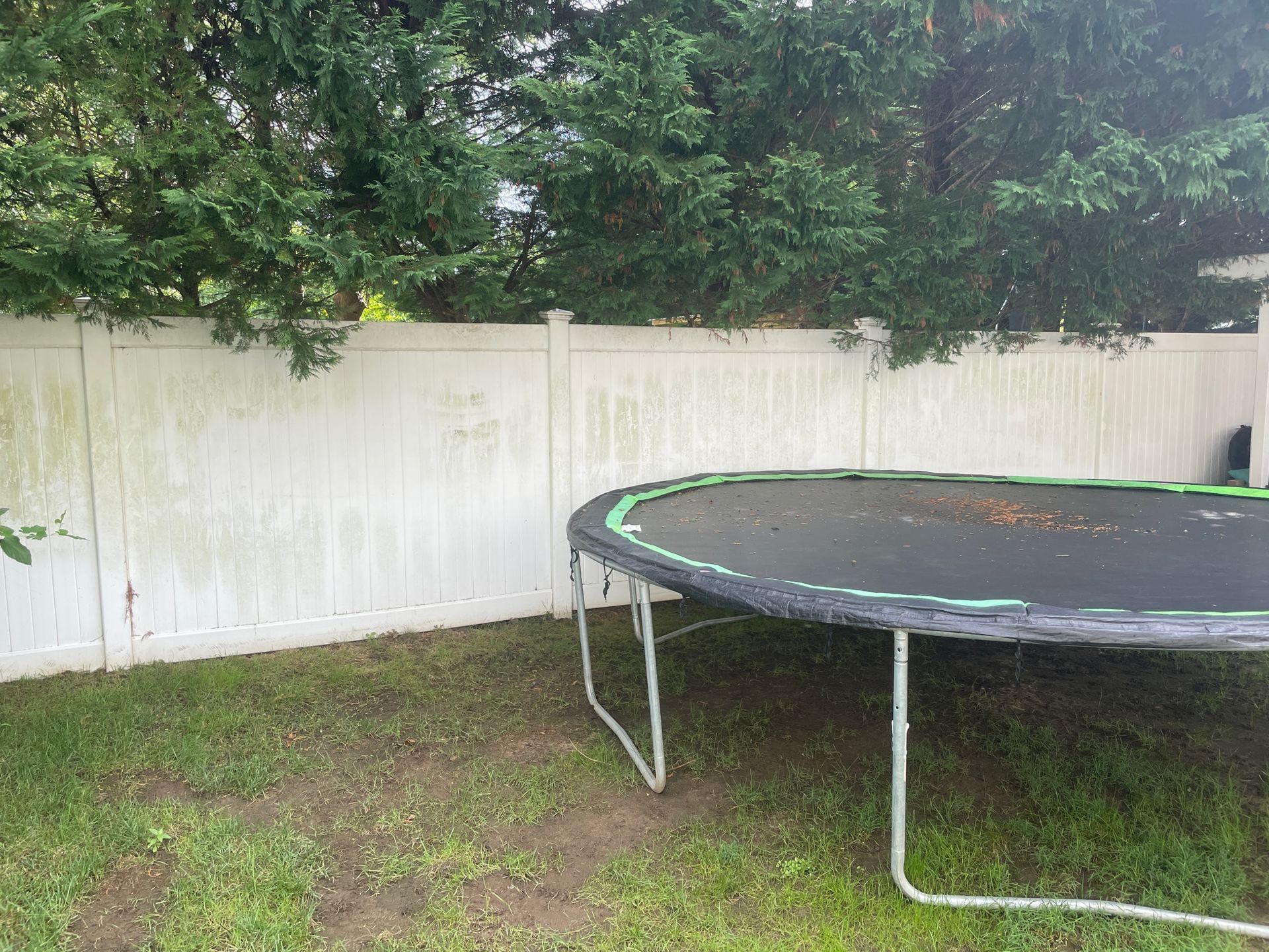 A backyard with a white fence, trampoline, and green grass. The fence has discoloration and some trees before.