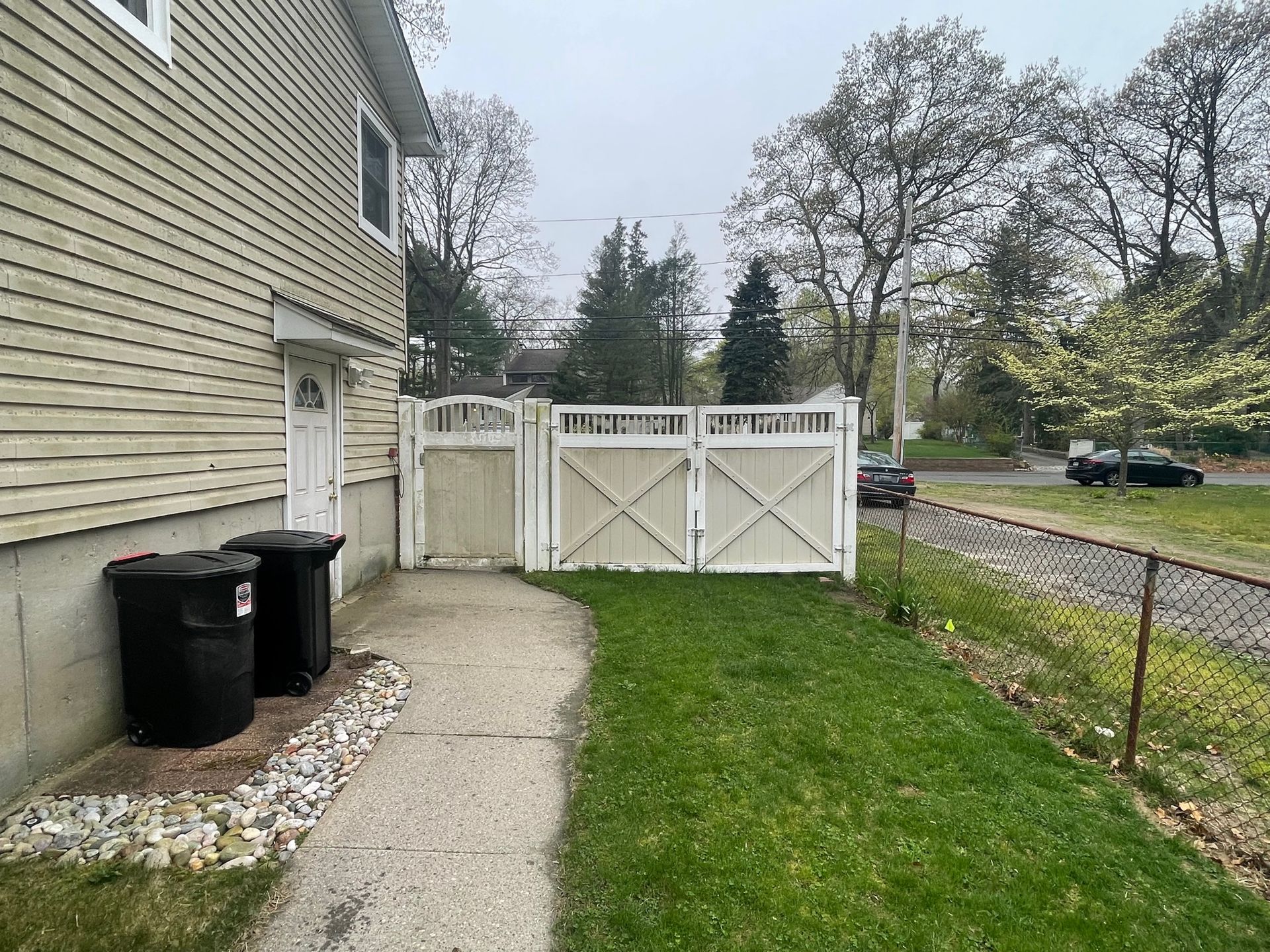 Back of a beige house with a white picket fence, two trash cans, and a grassy yard before.