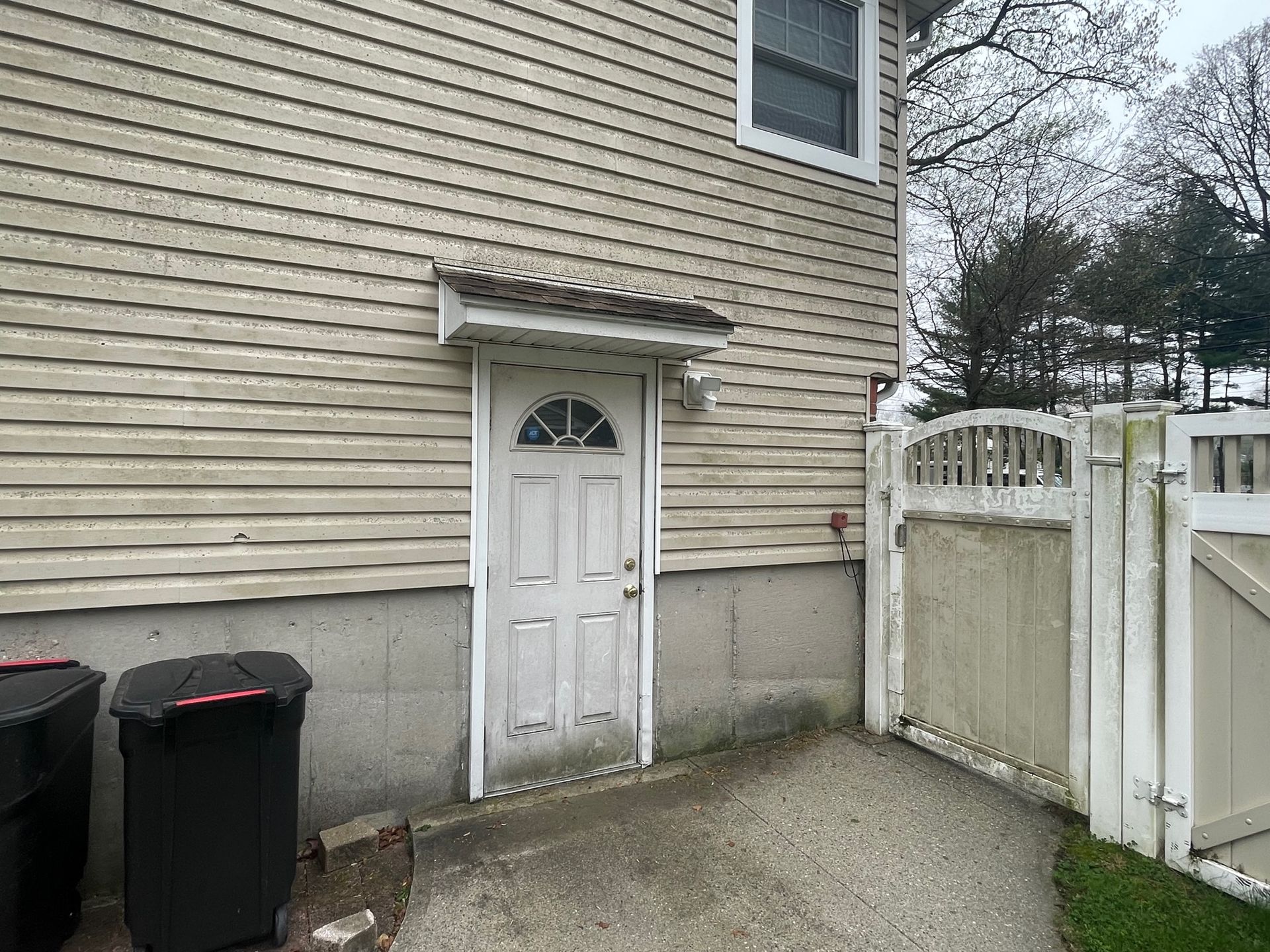 Back door of a house with a small awning, next to a white fence and trash cans before.