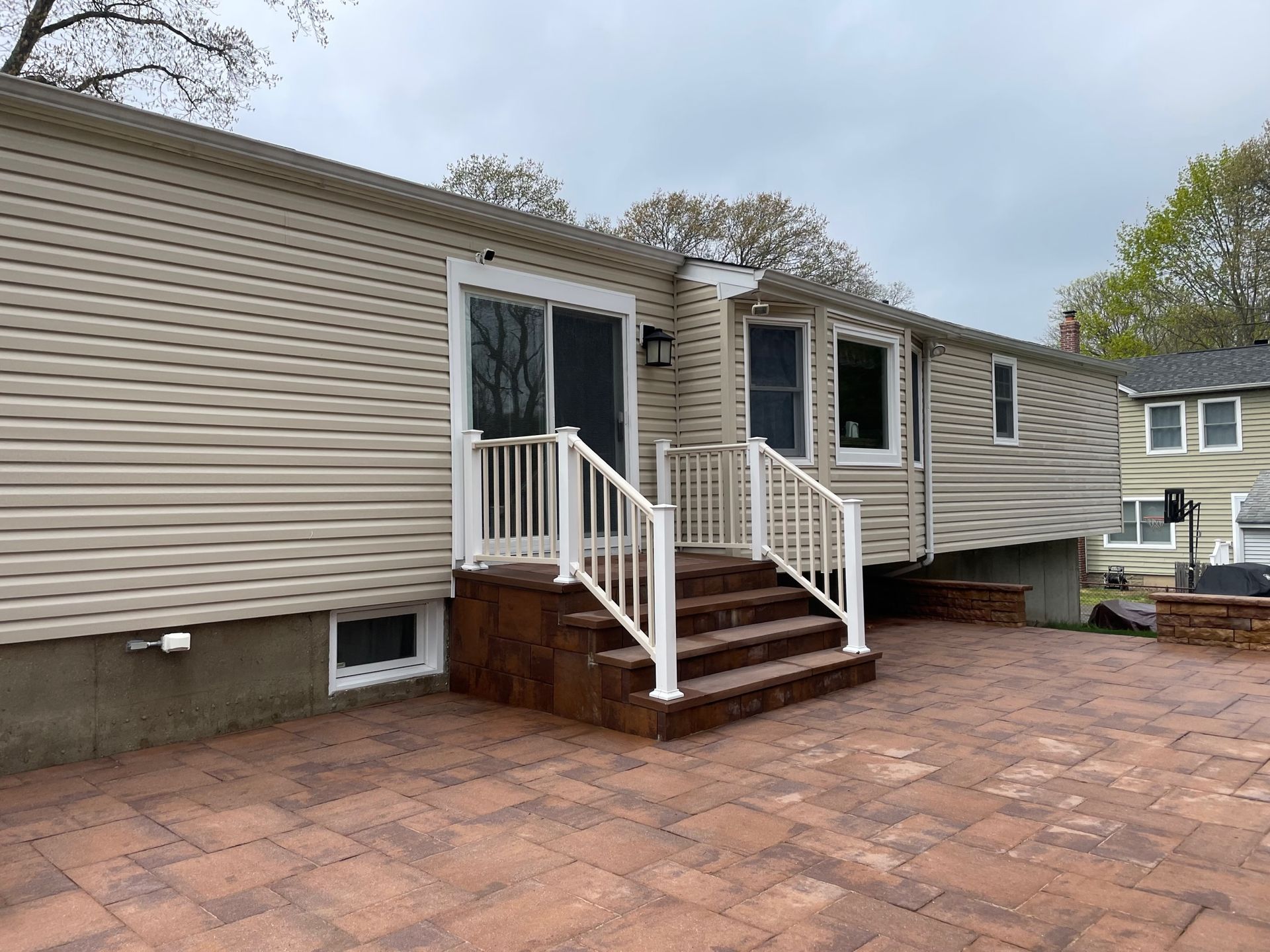 Back of house with brick patio, steps, and sliding door. Beige siding, white railing, overcast sky before.