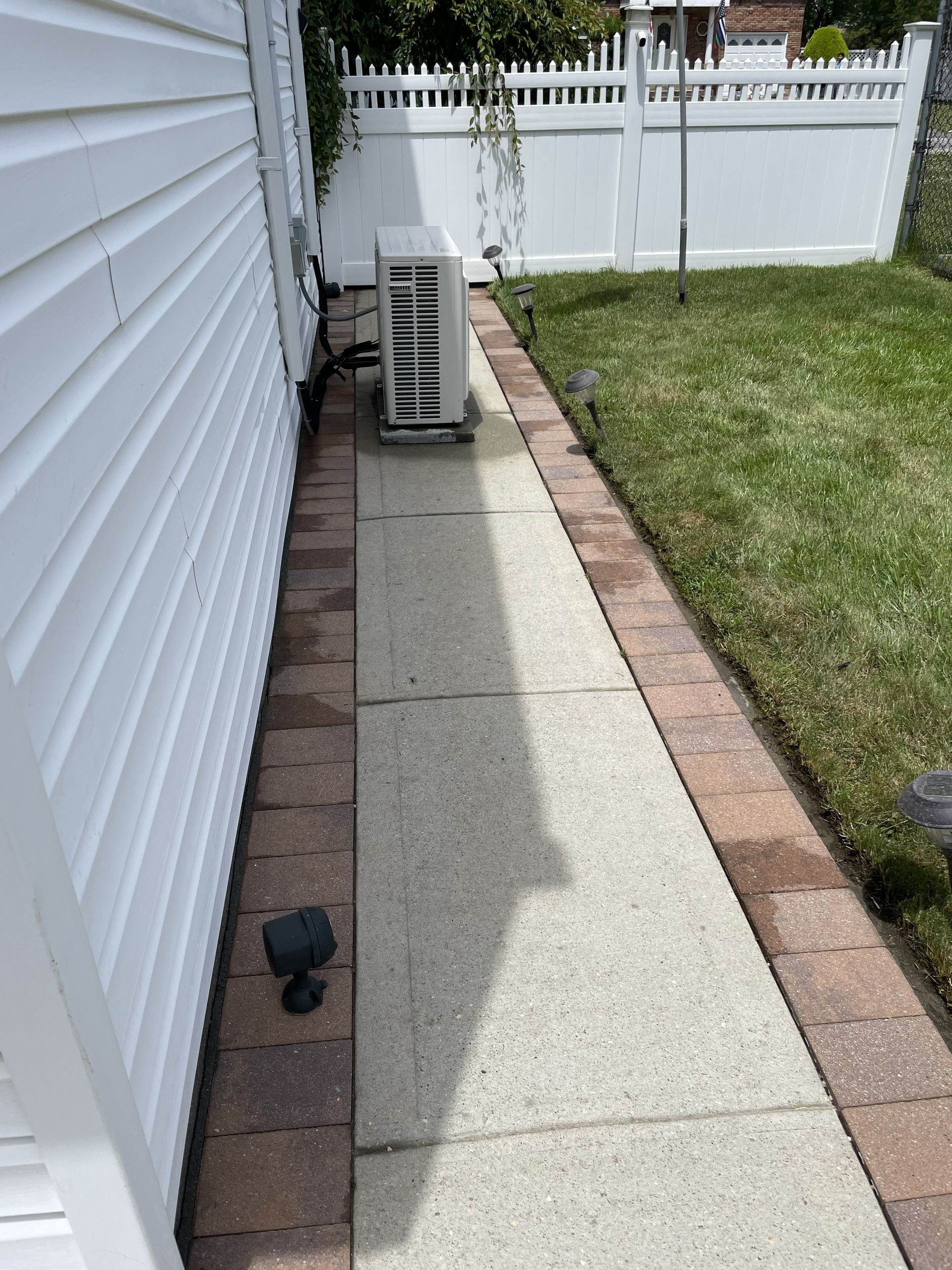 Narrow concrete side yard walkway beside white house, with outdoor AC unit and brick edging leading to a fence.