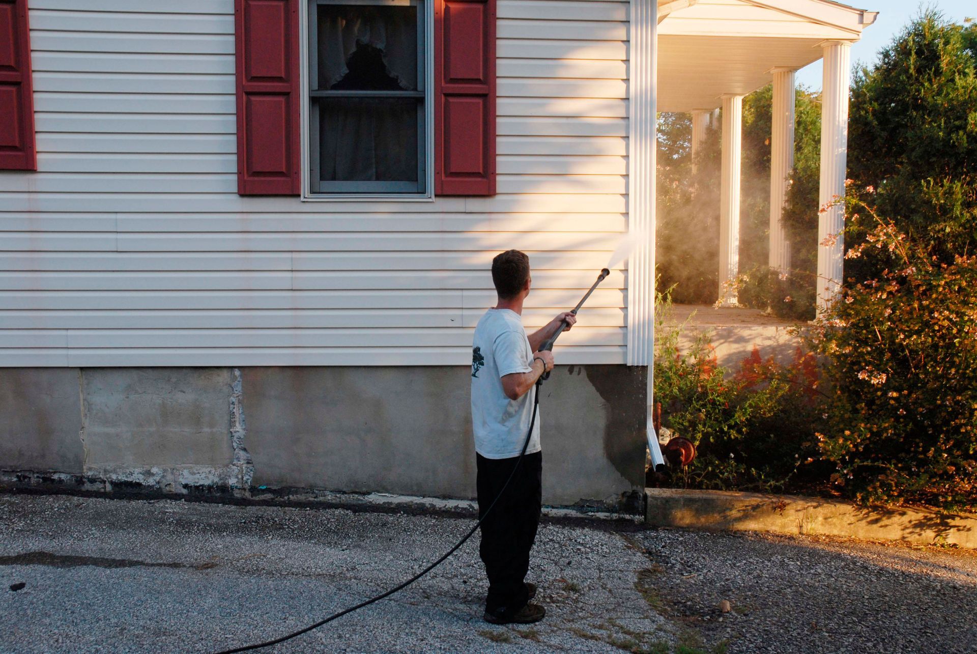 Person power washing the side of a white house with red shutters, spraying water towards a porch.
