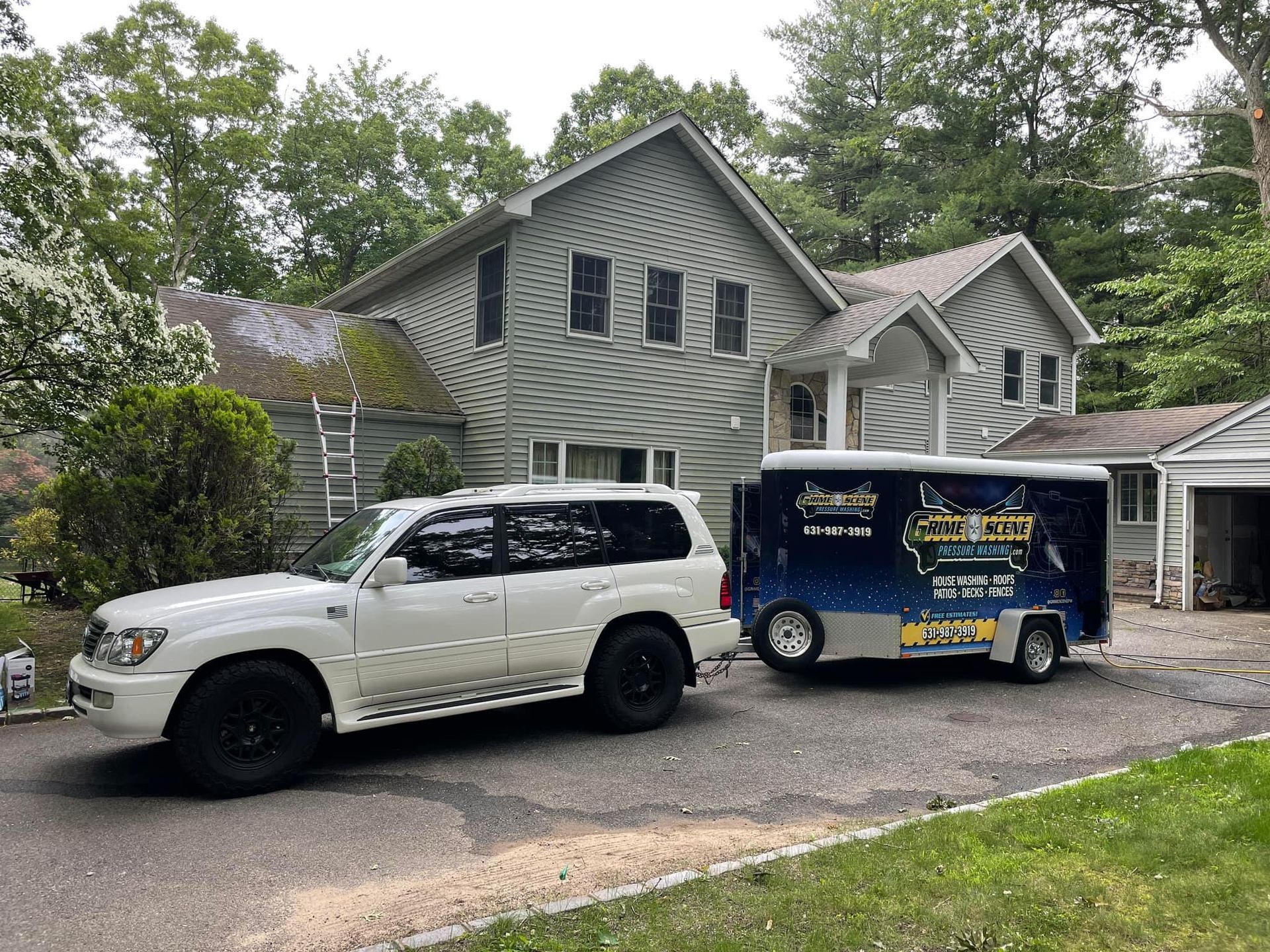 White SUV towing a trailer parked in front of a two-story gray house; trees in the background.