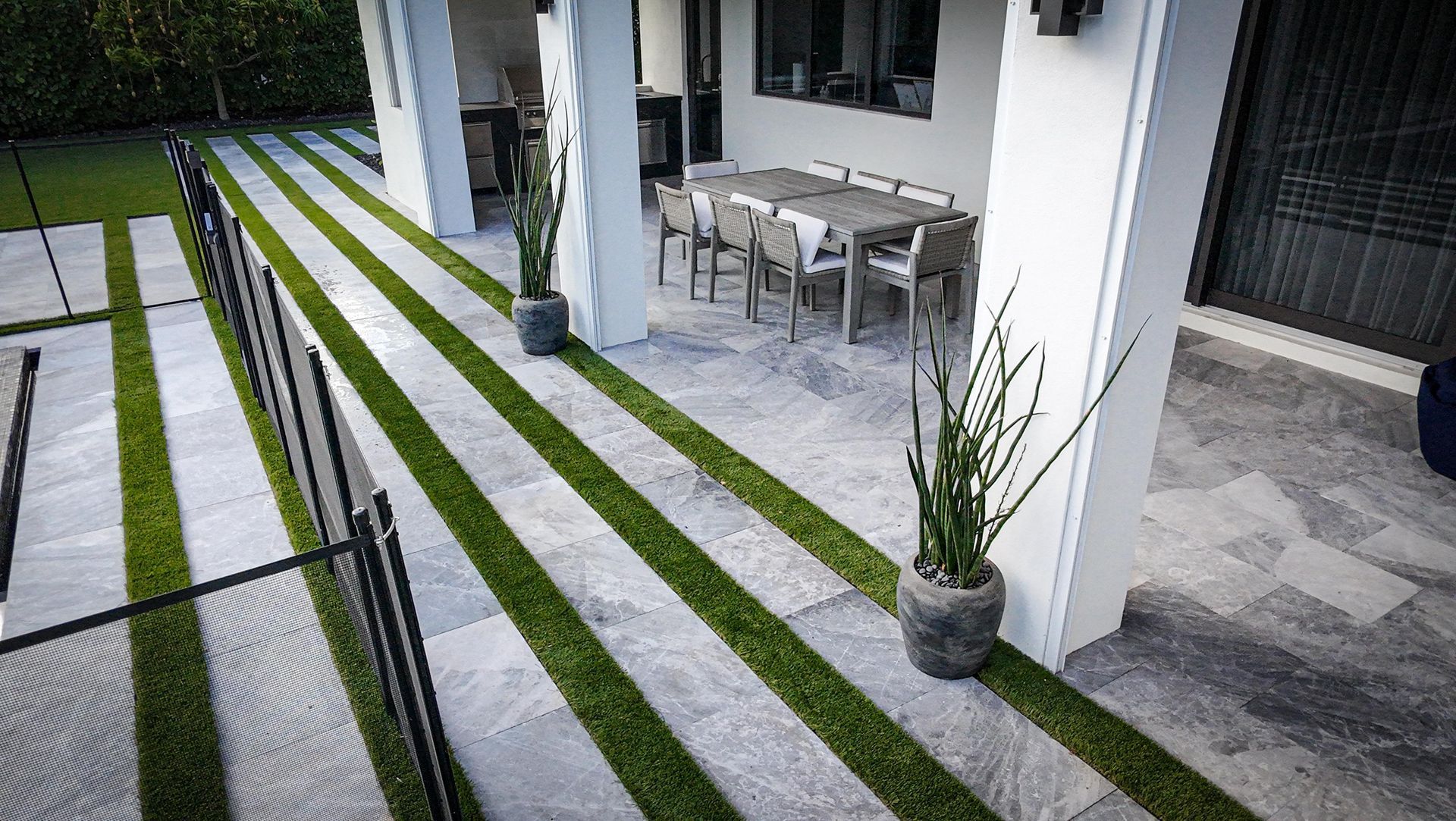 A patio with gray stone tiles featuring strips of green grass, outdoor dining furniture, and potted plants.