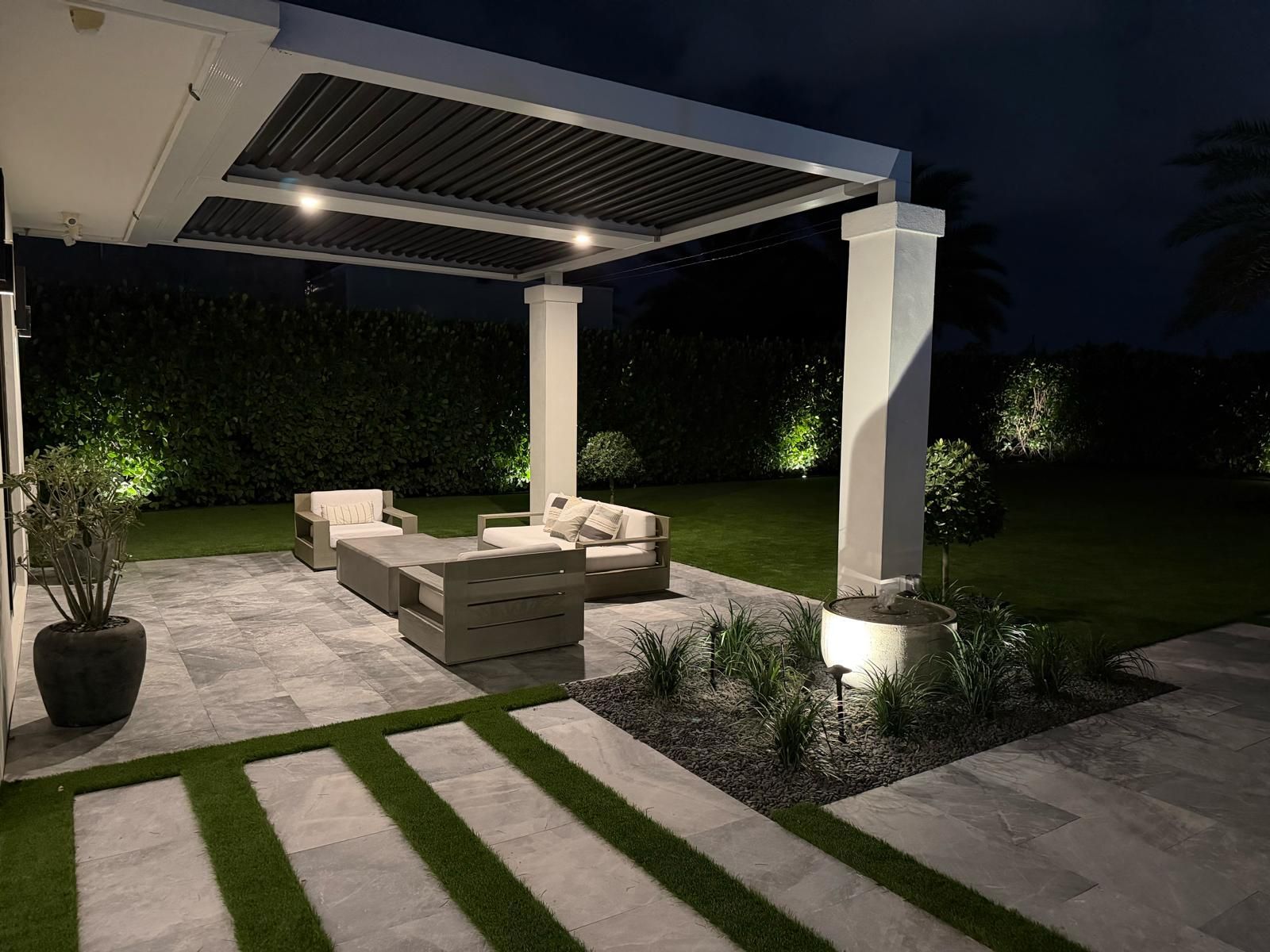 A patio at night with a pergola, gray outdoor furniture, lit landscaping, and stone pavers with grass strips.