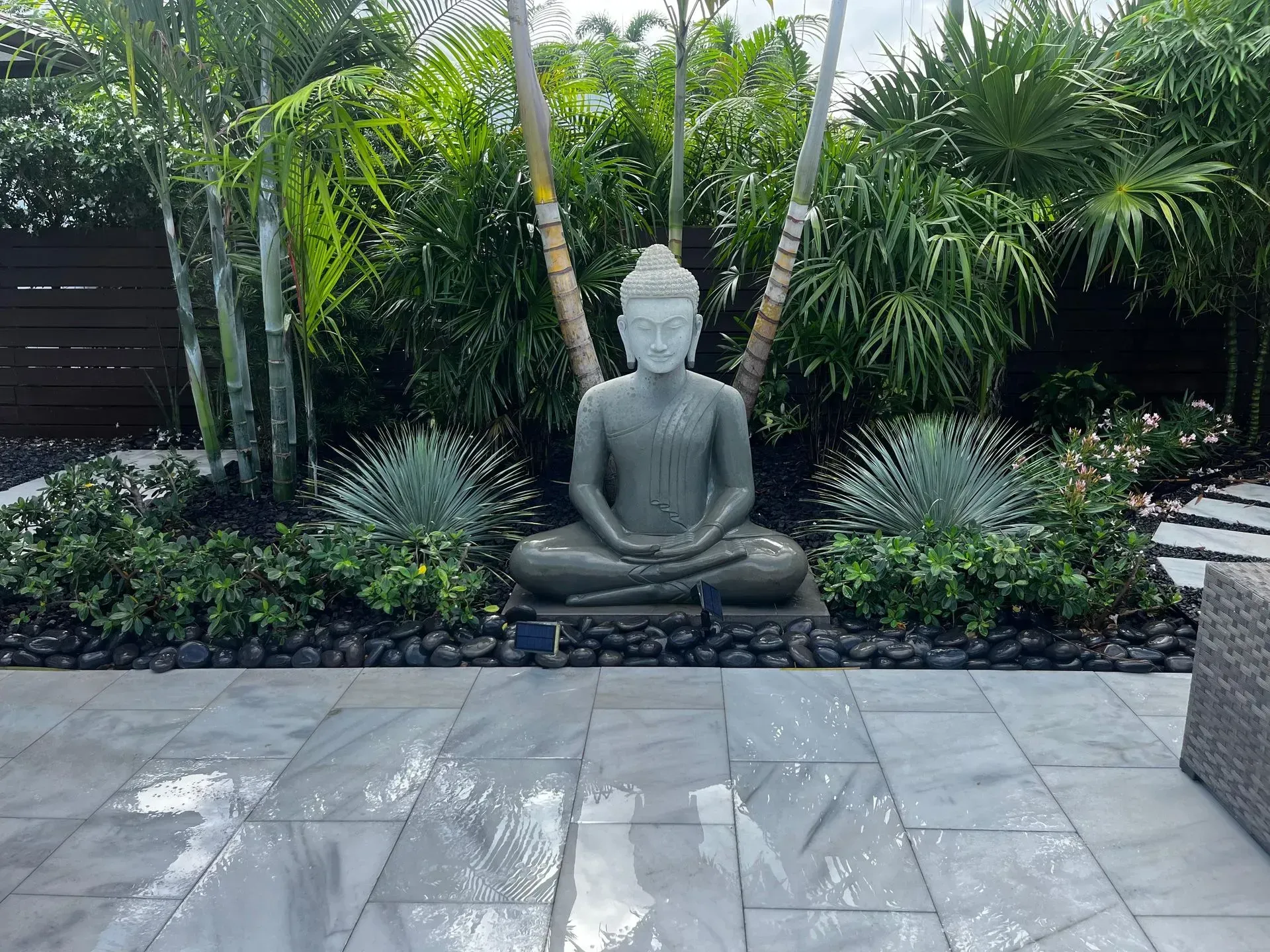 Stone Buddha statue in a lush garden setting with palm trees and a tiled patio.