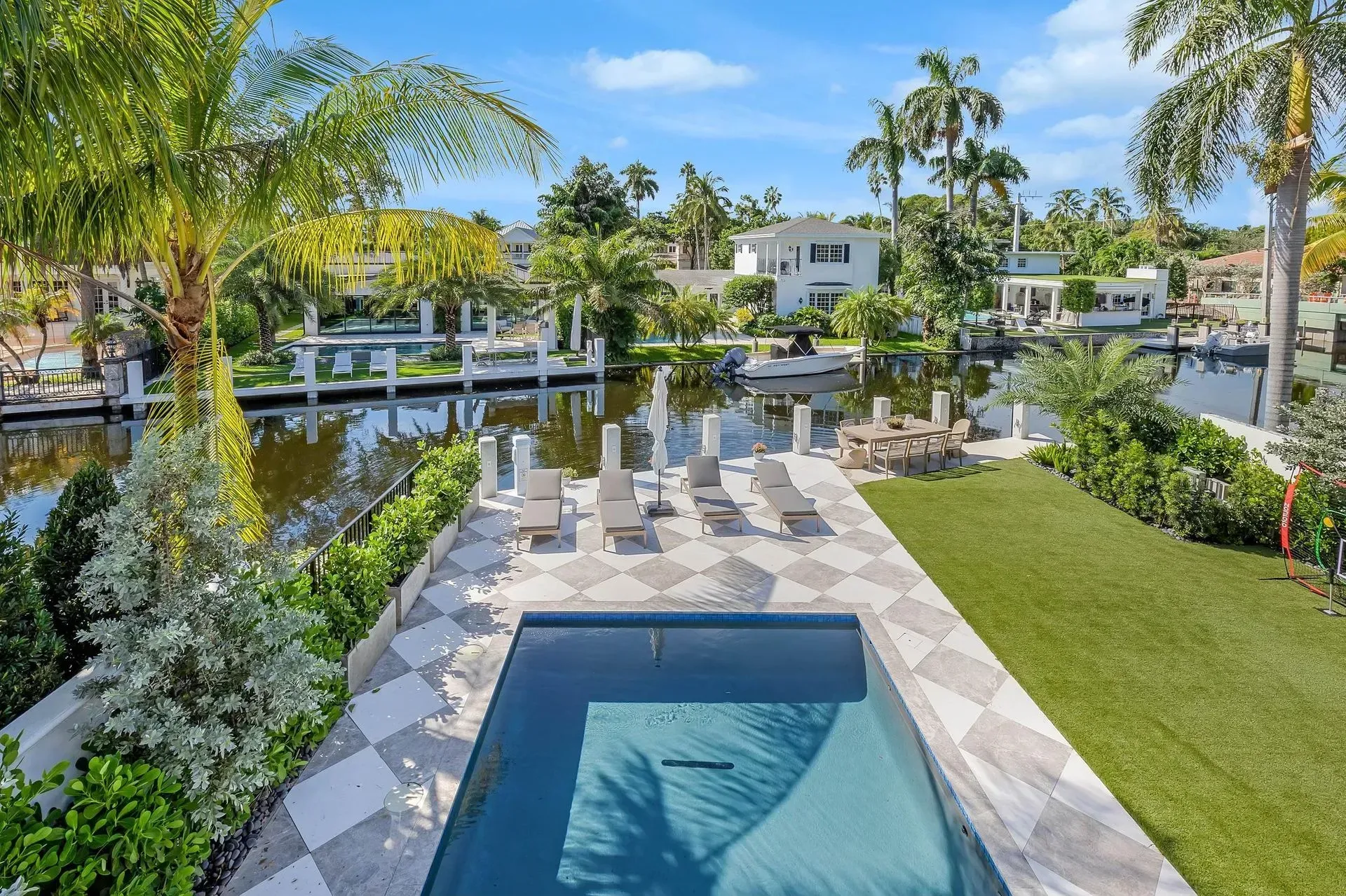 Pool overlooking canal with lounge chairs, grass, palm trees, and white houses.