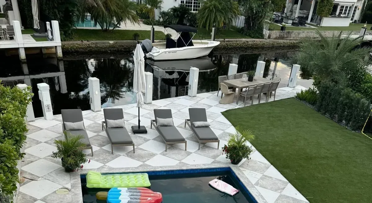 Poolside patio with pool, chaise lounges, and dining table, next to a canal with a boat.