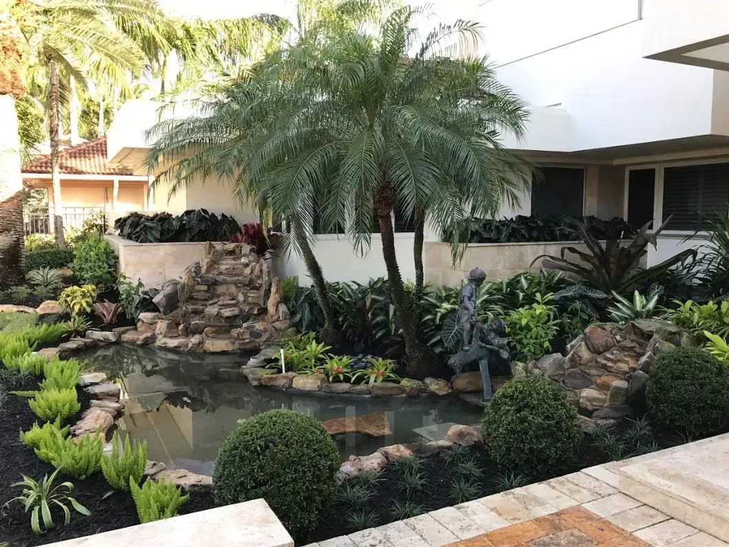 Landscaped garden with pond, palm tree, statue, and lush greenery. White building in the background.