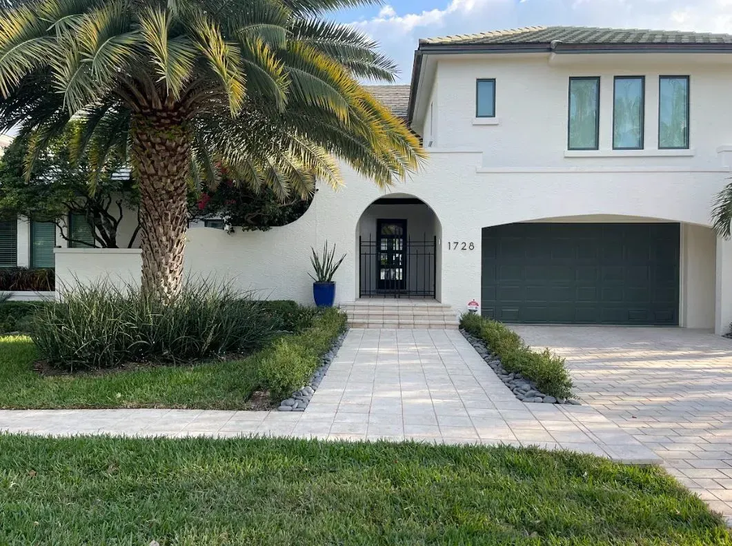 White house with arched entryway, dark green garage door, palm tree, and brick walkway.
