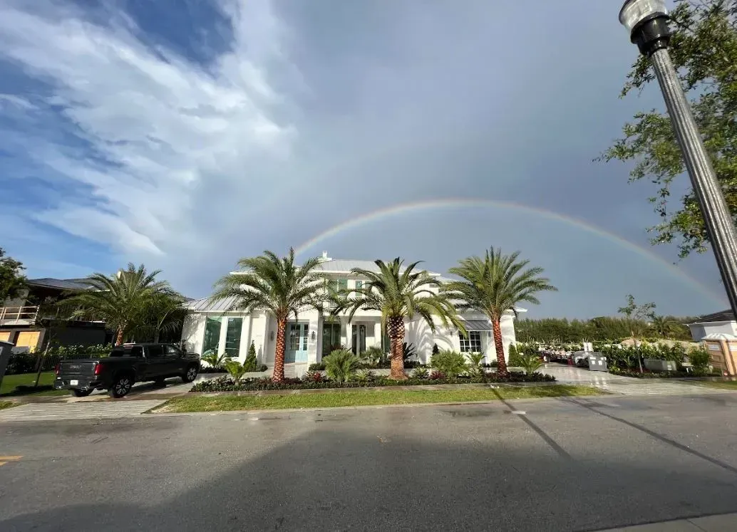 Rainbow arcs over a white house with palm trees, blue sky with clouds. A street is in the foreground.