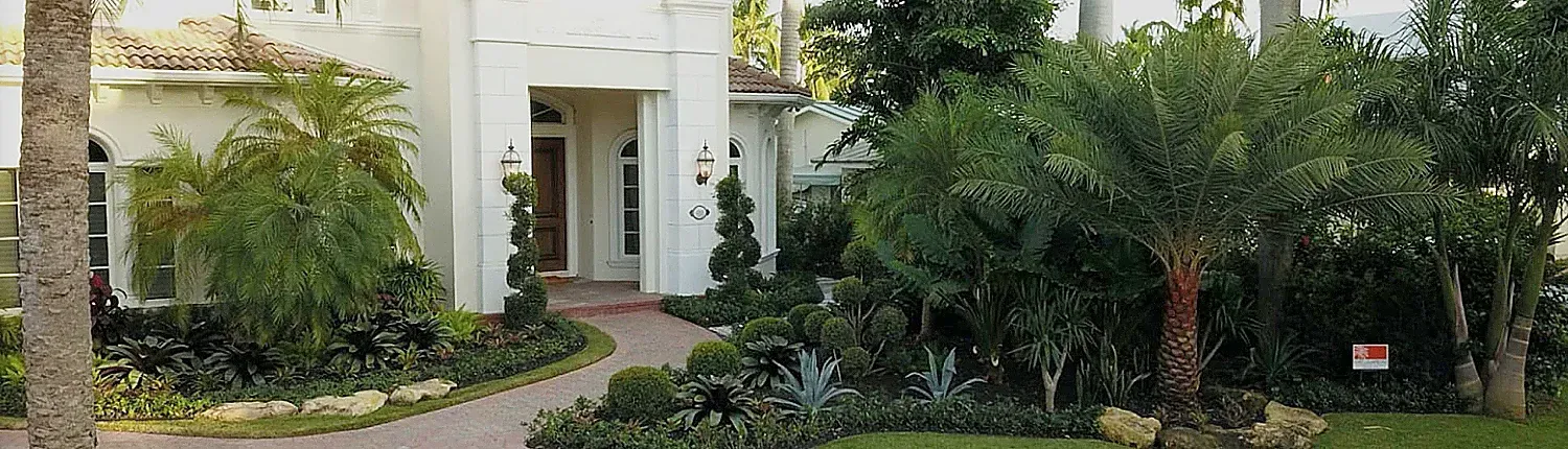 Exterior view of a white stucco house with a brick pathway leading to the front door, surrounded by lush green foliage.