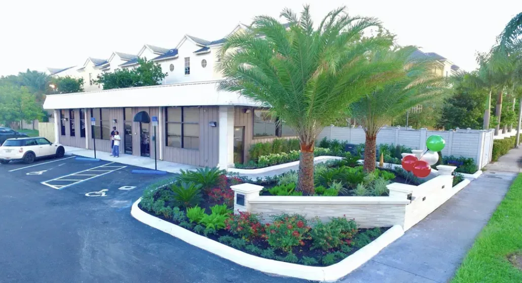 Exterior view of a one-story building with a landscaped garden and palm trees; a car is parked in front.