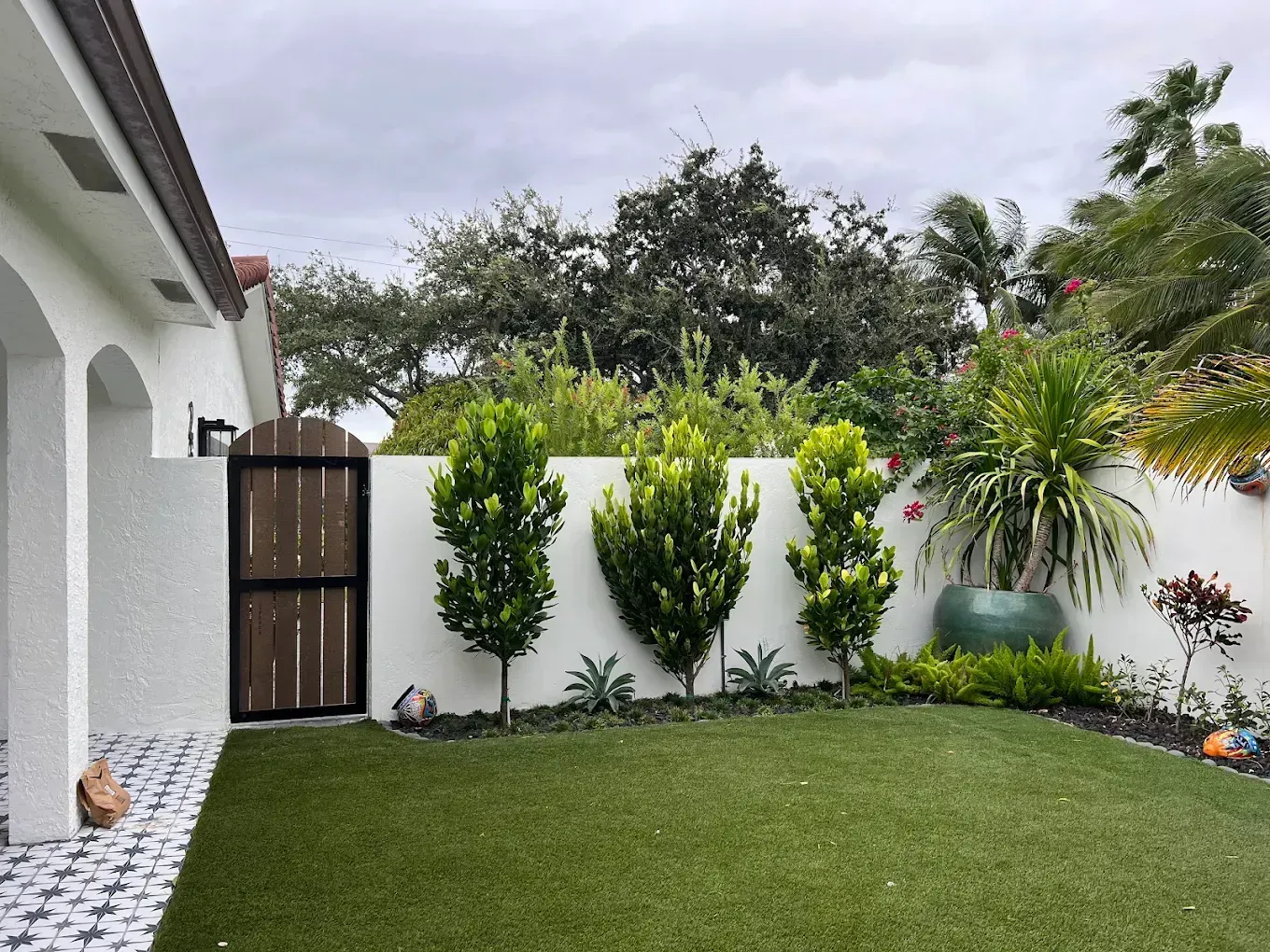 A small backyard with a faux grass lawn, white walls, wooden gate, and a line of green trees against a cloudy sky.