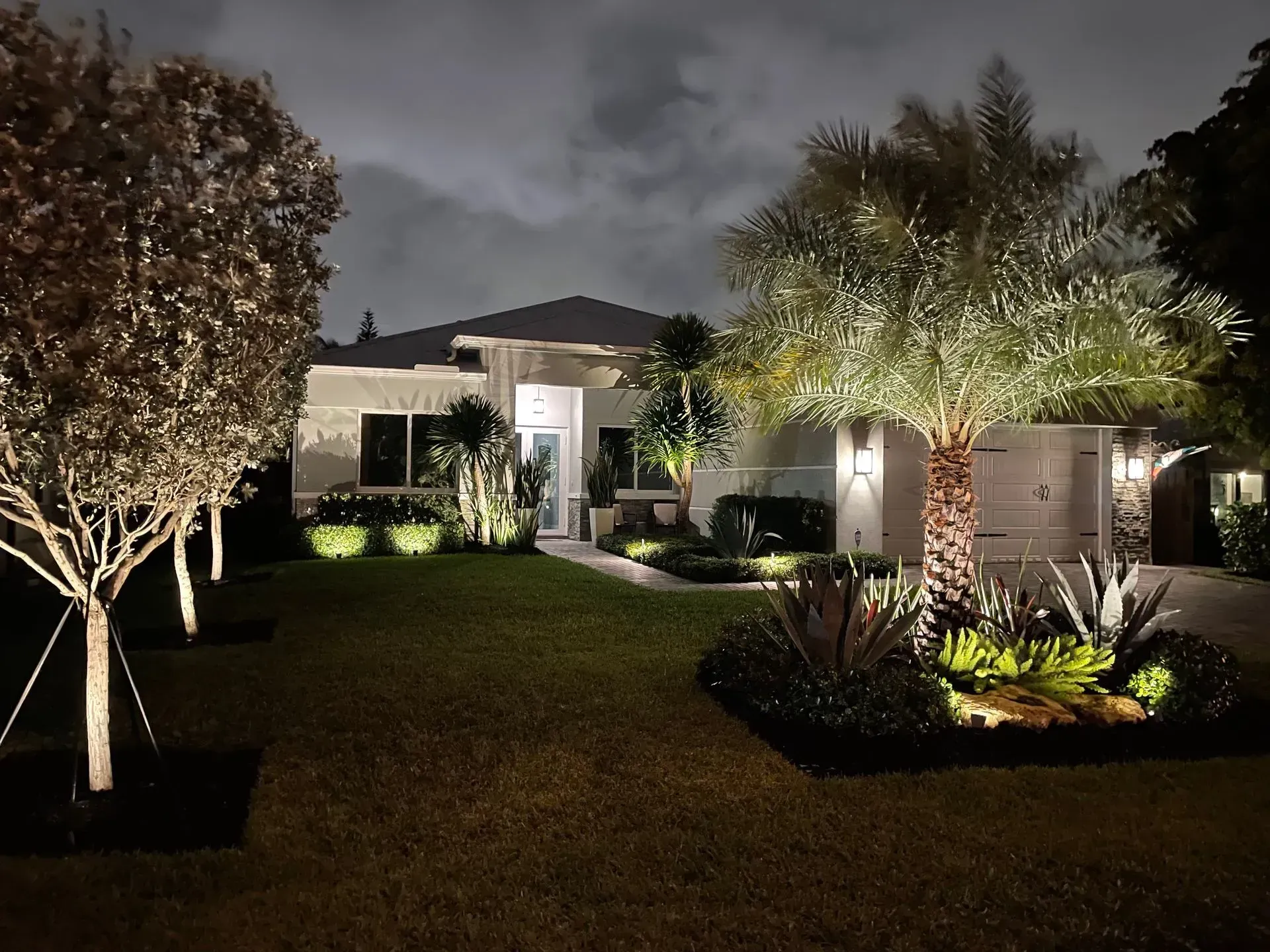 A well-lit house at night with trees and landscaping.