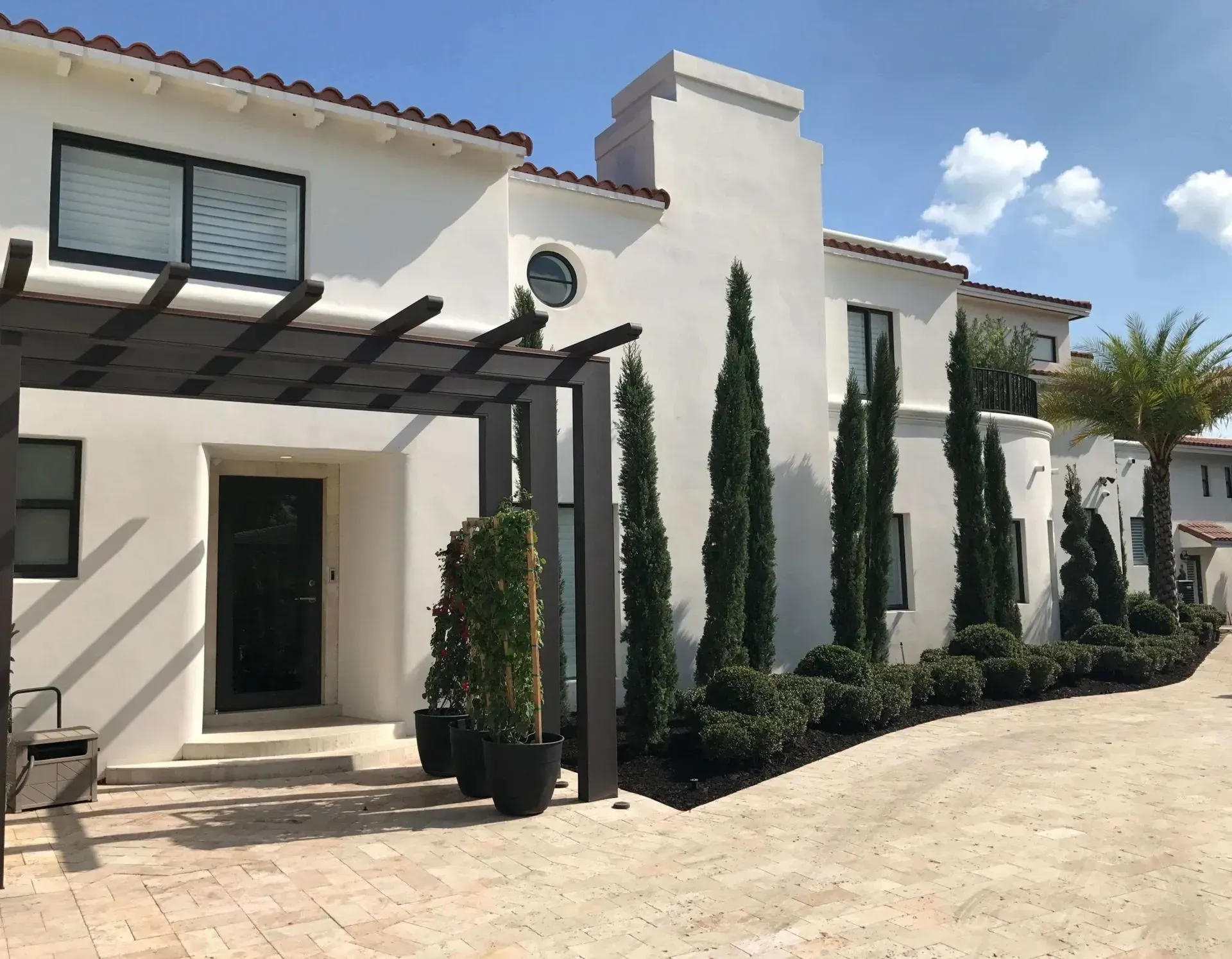 White stucco building with red tile roof, cypress trees, and a pergola.