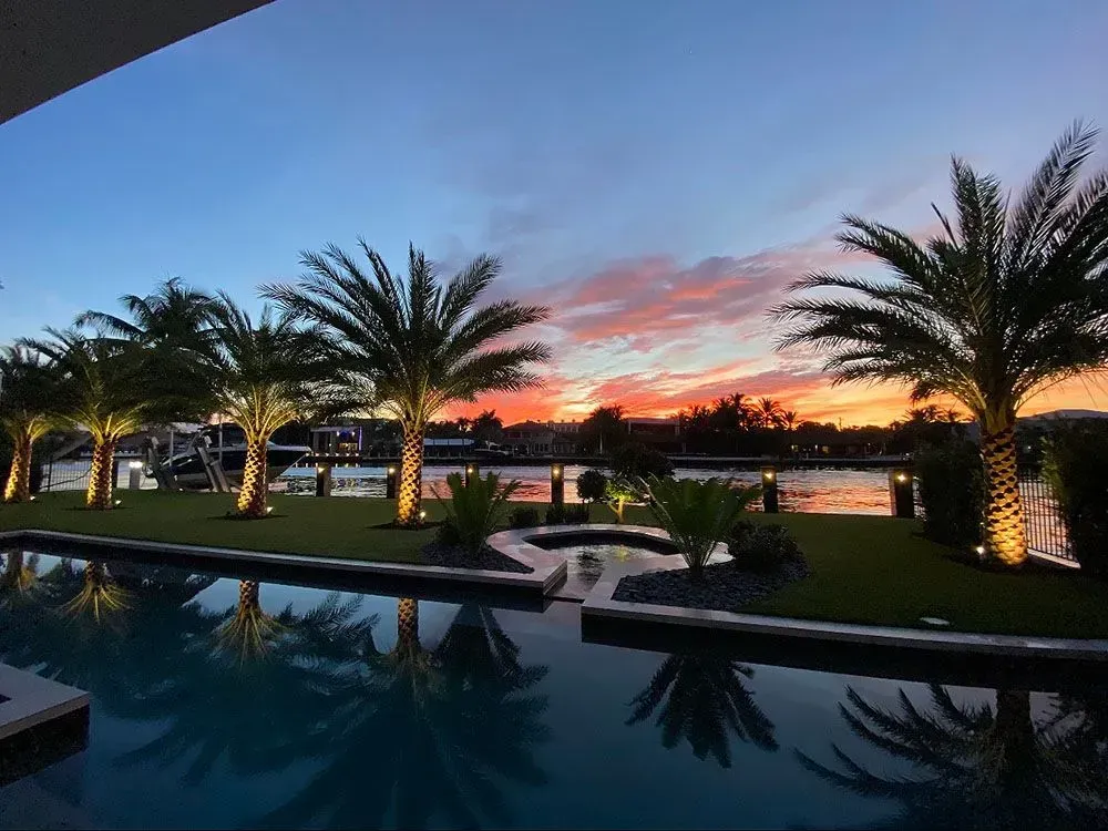 Palm trees line a canal, reflecting in a pool at sunset with orange and pink hues.