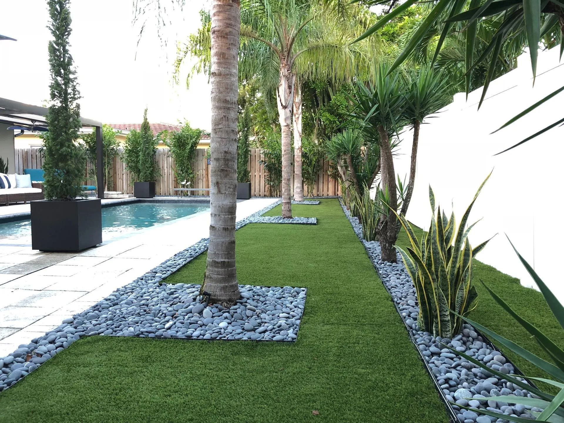 Backyard with pool, palm trees, and a strip of green turf bordered by grey stones against a white fence.